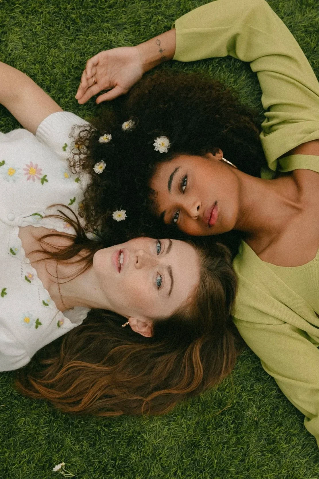 Deux femmes allongées sur de l'herbe verte, l'une avec des cheveux bruns et l'autre avec des cheveux bouclés noirs, entourées de petites fleurs blanches dans leurs cheveux, regardant vers l'objectif.