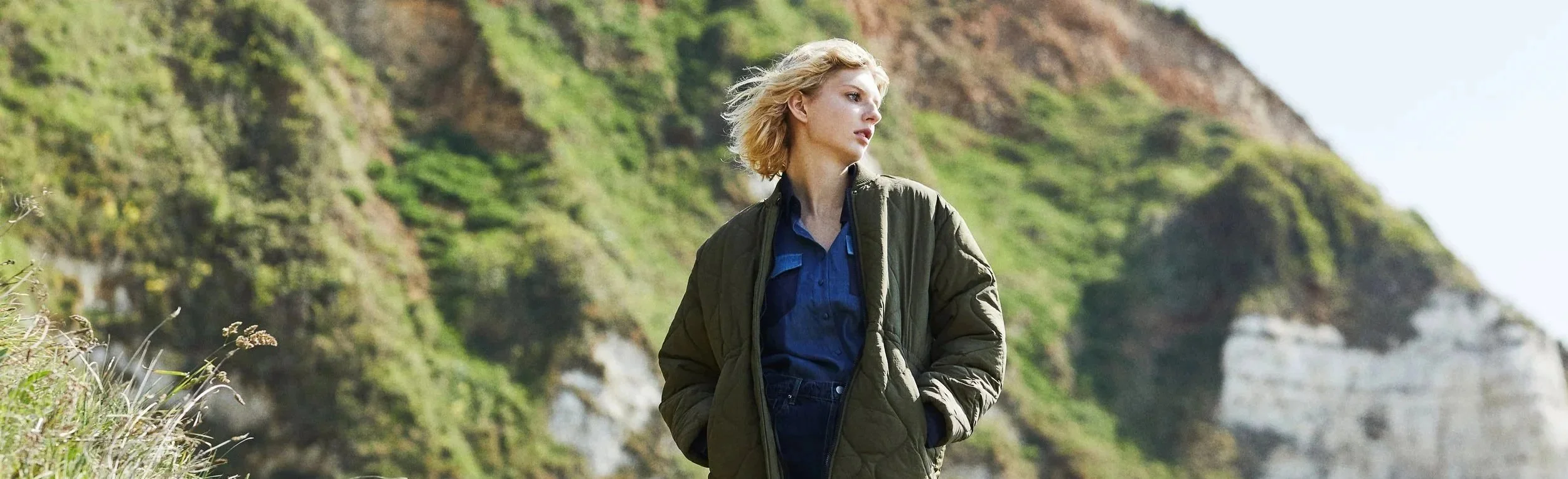 Une femme blonde regarde vers la droite, portant une veste kaki et un jean, avec un paysage de falaises verdoyantes en arrière-plan.