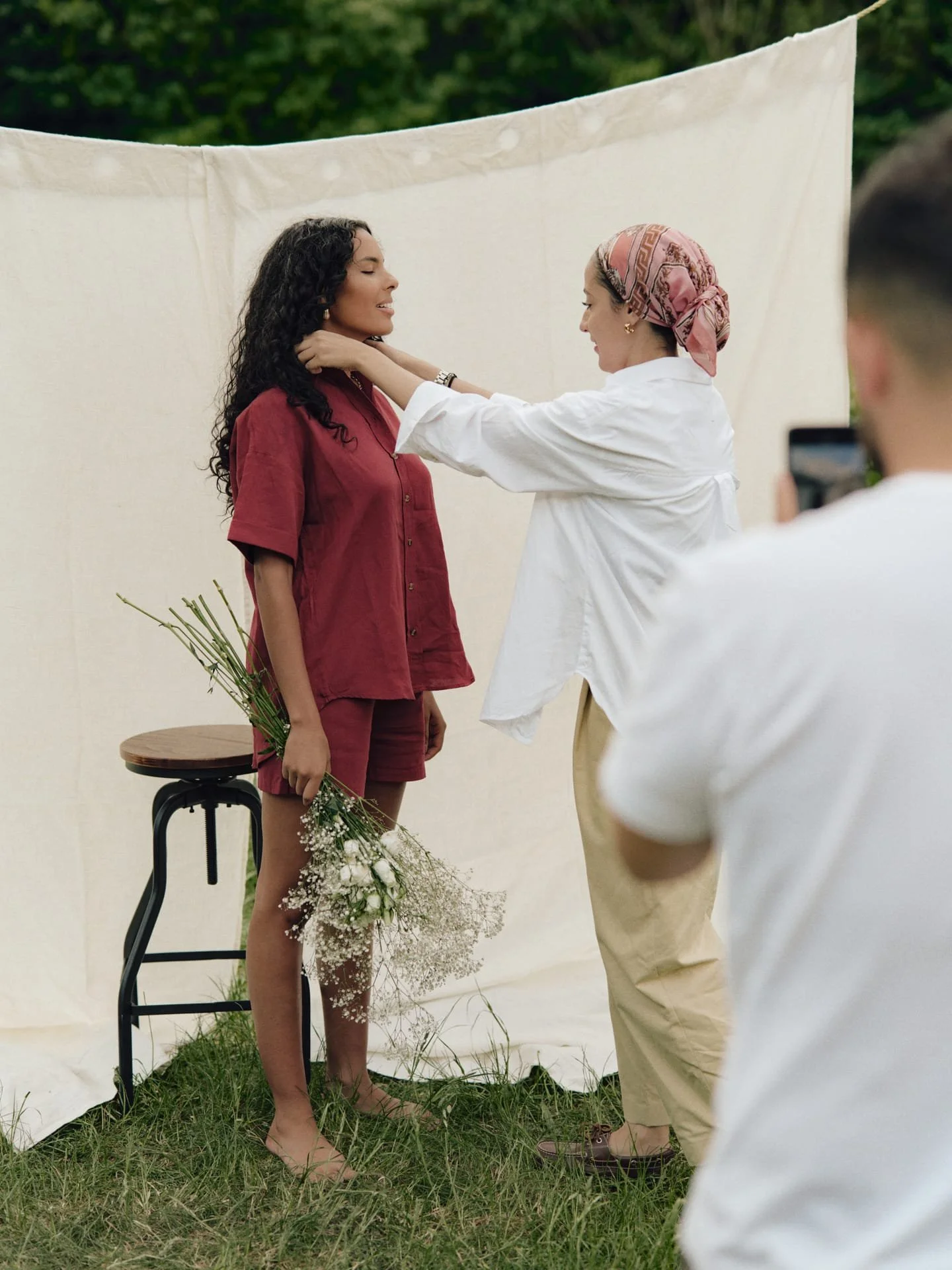 Une séance de photographie avec deux femmes, une debout avec un bouquet de fleurs, l'autre ajustant le col de la première, sous un drap blanc en plein air.