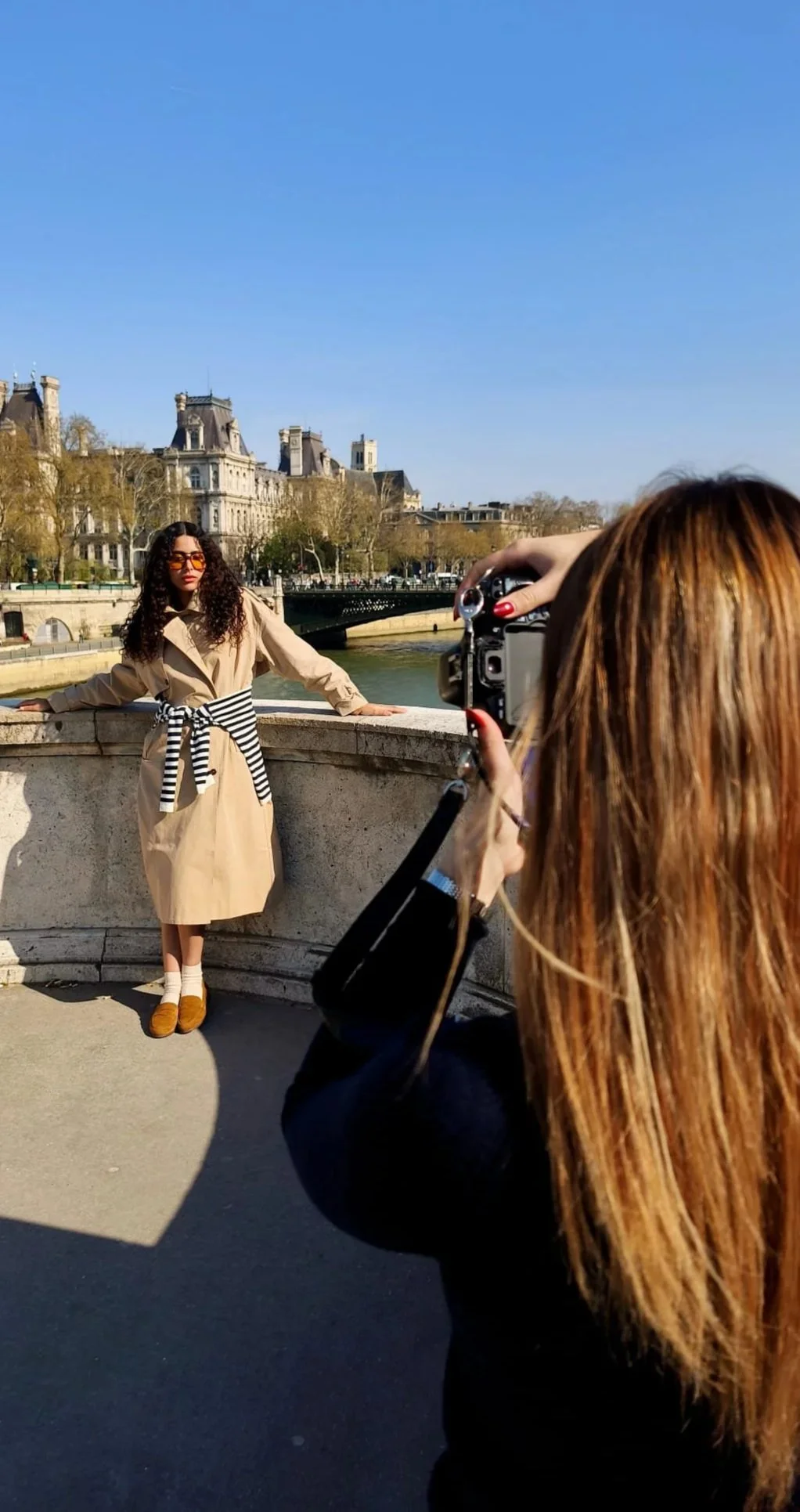 Une femme avec des cheveux bouclés bruns foncés pose pour une photo devant la Seine à Paris, portant une trench-coat beige, des chaussettes blanches, des mocassins marron, et des lunettes de soleil orange, avec la Tour Eiffel en fond.