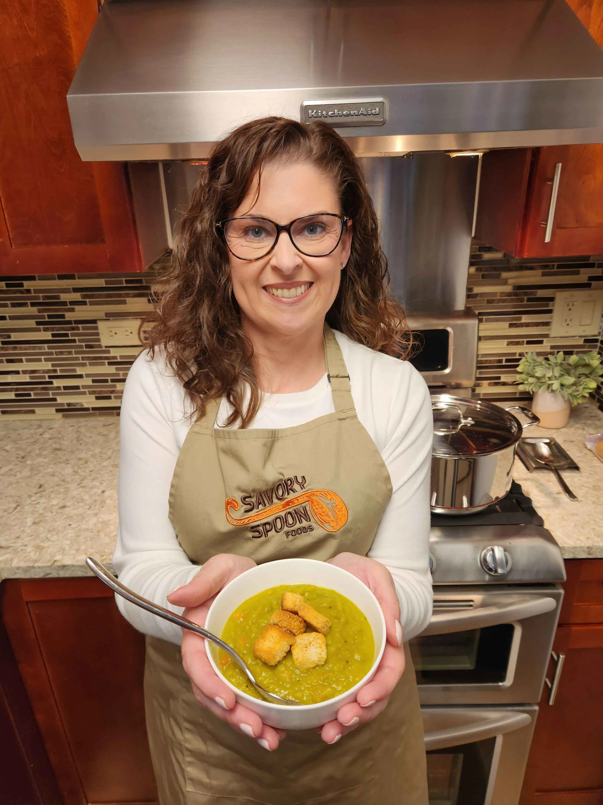 Owner Deanna Fritzinger, a woman with curly brown hair and glasses smiling, wearing an apron, holding a bowl of their split pea and ham soup with croutons in a kitchen.