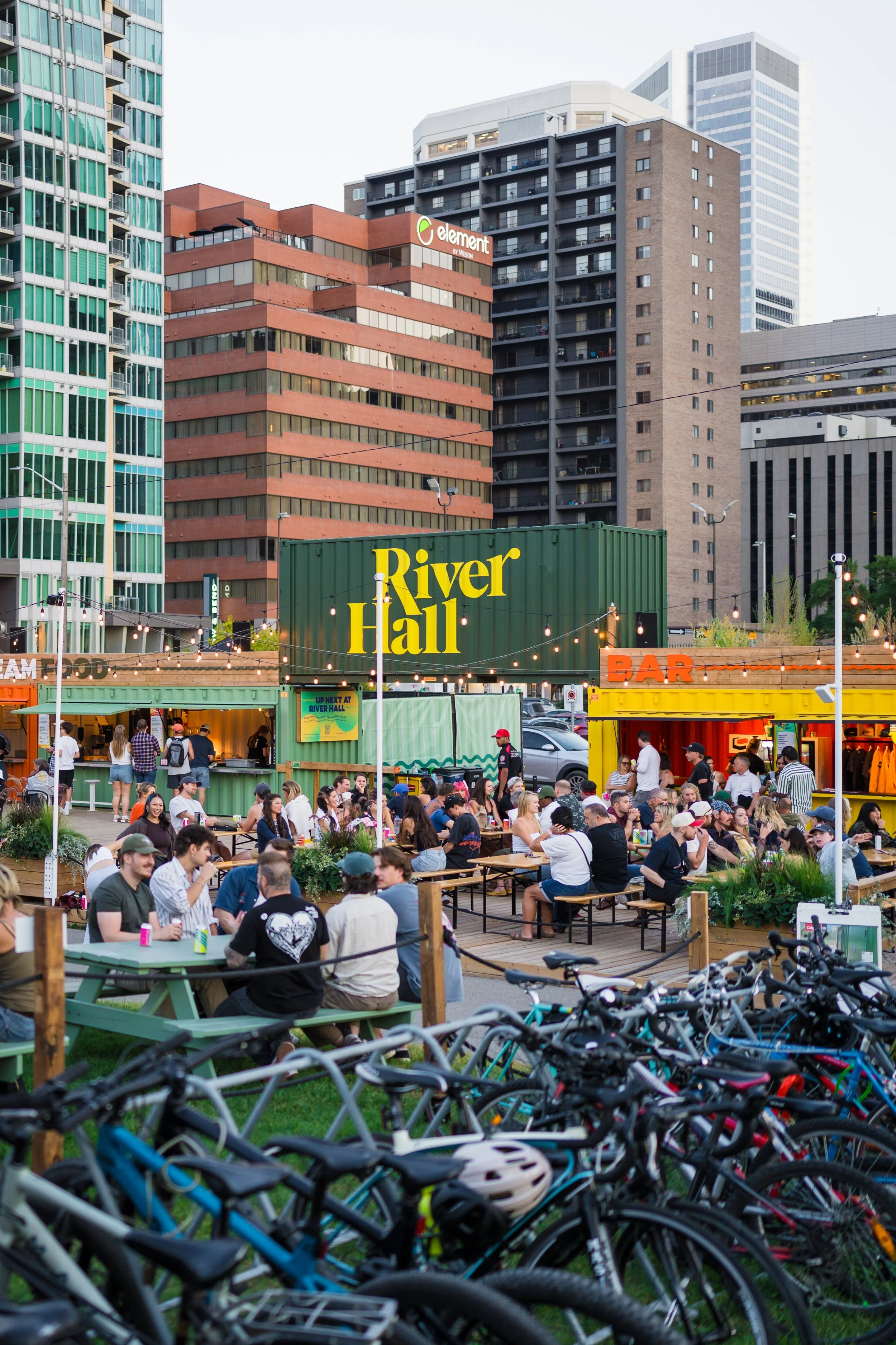 River Hall's deck on a sunny day filled with people sitting at picnic tables, surrounded by bicycles, with a cityscape of tall buildings in the background. There are food and bar stalls, and signs reading "River Hall" and "BAR."