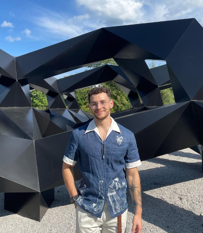 Young man with glasses smiling outdoors in front of a modern, geometric black sculpture with a tunnel-like structure. He is wearing a denim shirt with white accents and illustrations, white pants, and a watch.