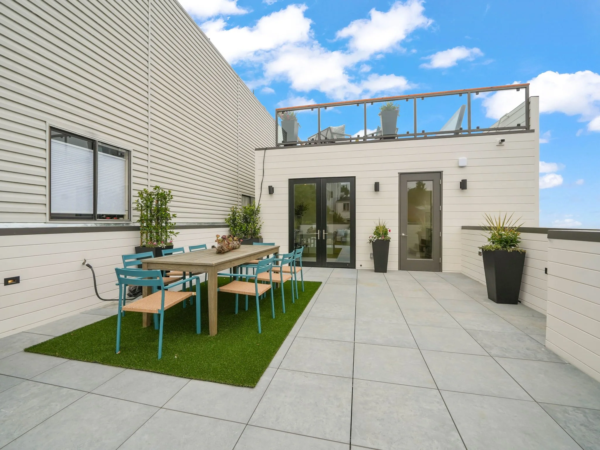 Modern outdoor patio with a dining table and teal chairs, potted plants, large beige building walls, glass door and window, blue sky with clouds.