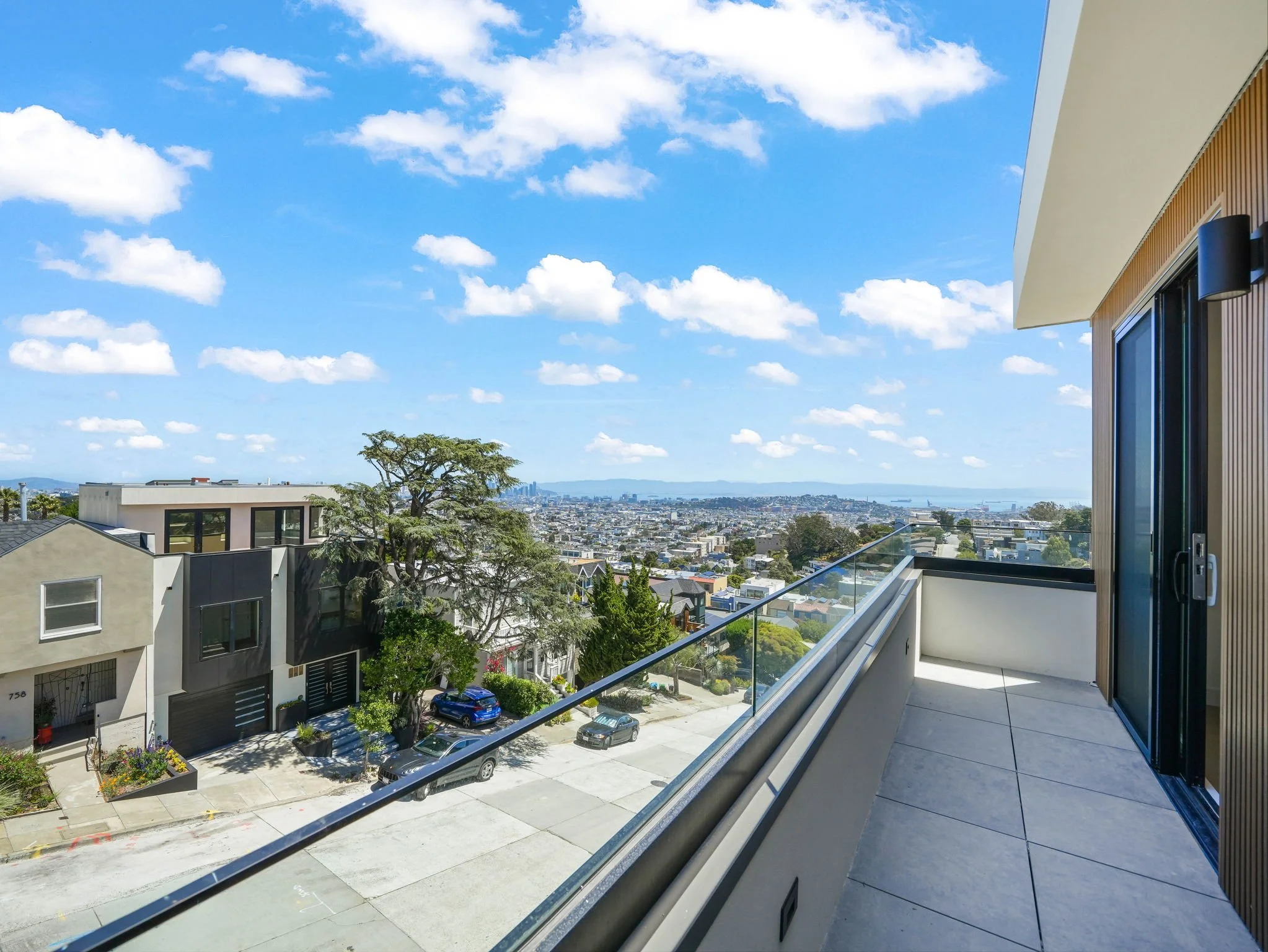 View from a balcony showing a cityscape, residential buildings, a tree, parking lot, and a distant body of water under a blue sky with scattered clouds.