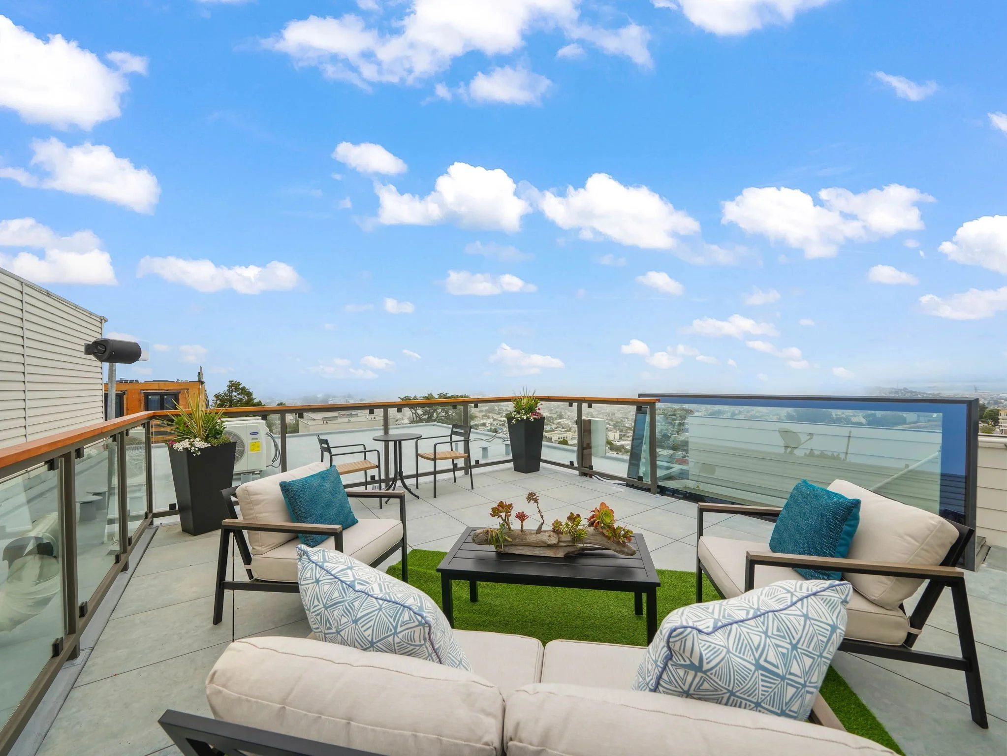 A rooftop patio with outdoor furniture, including cream-colored sofas with striped cushions, black metal chairs with blue cushions, small tables, potted plants, and a scenic city view with a cloudy blue sky in the background.