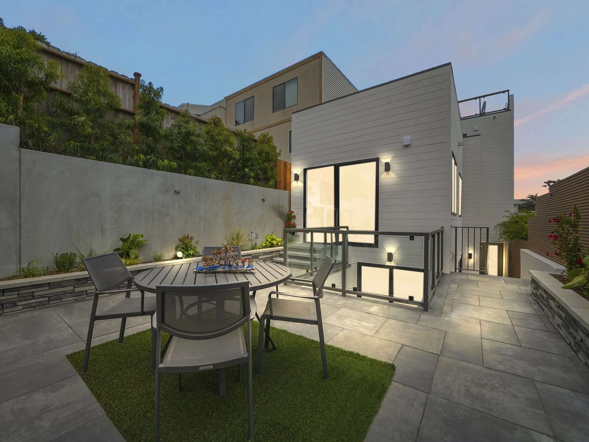Modern rooftop patio with outdoor dining table, chairs, and potted plants, overlooking neighboring buildings and a clear sky at dusk.