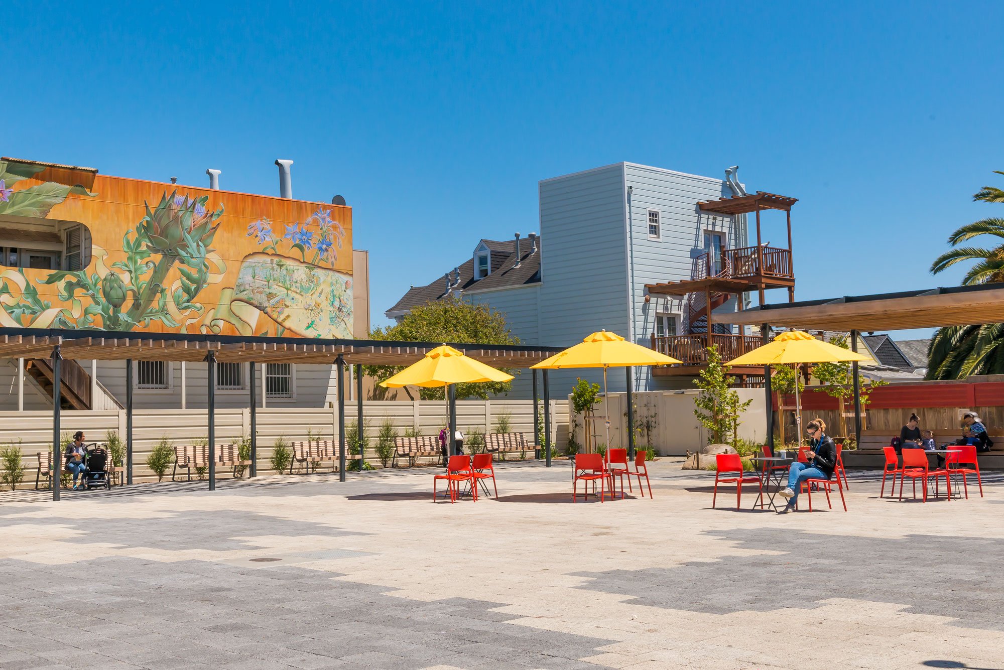 Outdoor plaza with red chairs, yellow umbrellas, mural, and blue sky