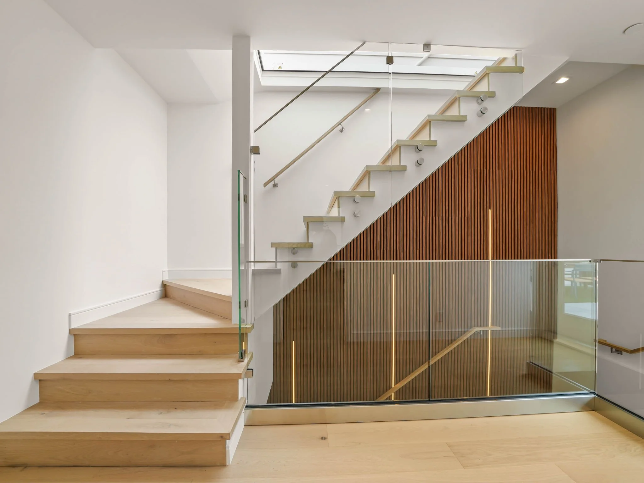 Interior view of a modern staircase with light wooden steps, glass railings, and wooden slat wall paneling.