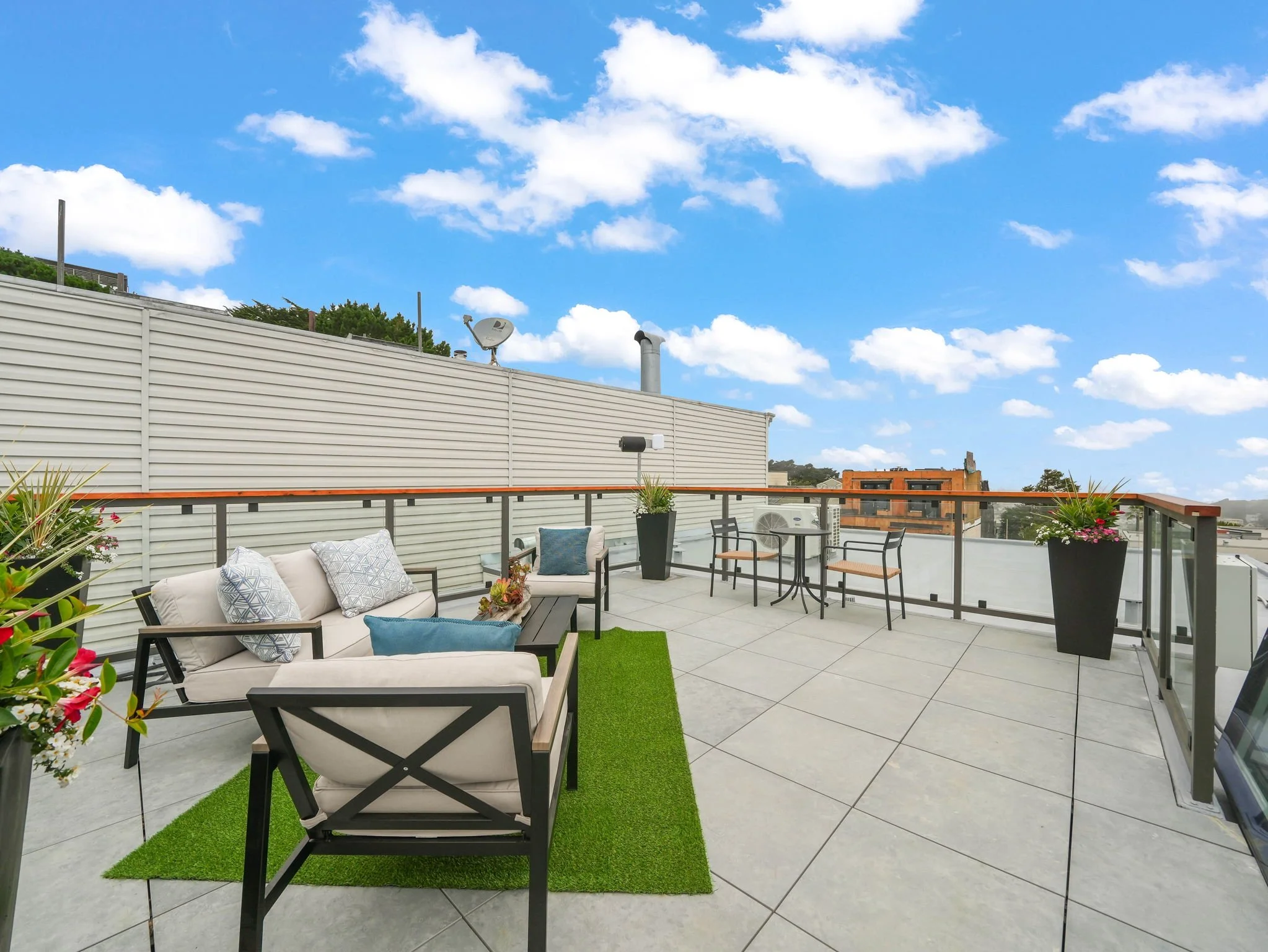 Rooftop patio with outdoor furniture including chairs and a sofa, potted plants, a green artificial turf runner, a railing, and a partly cloudy sky in the background.
