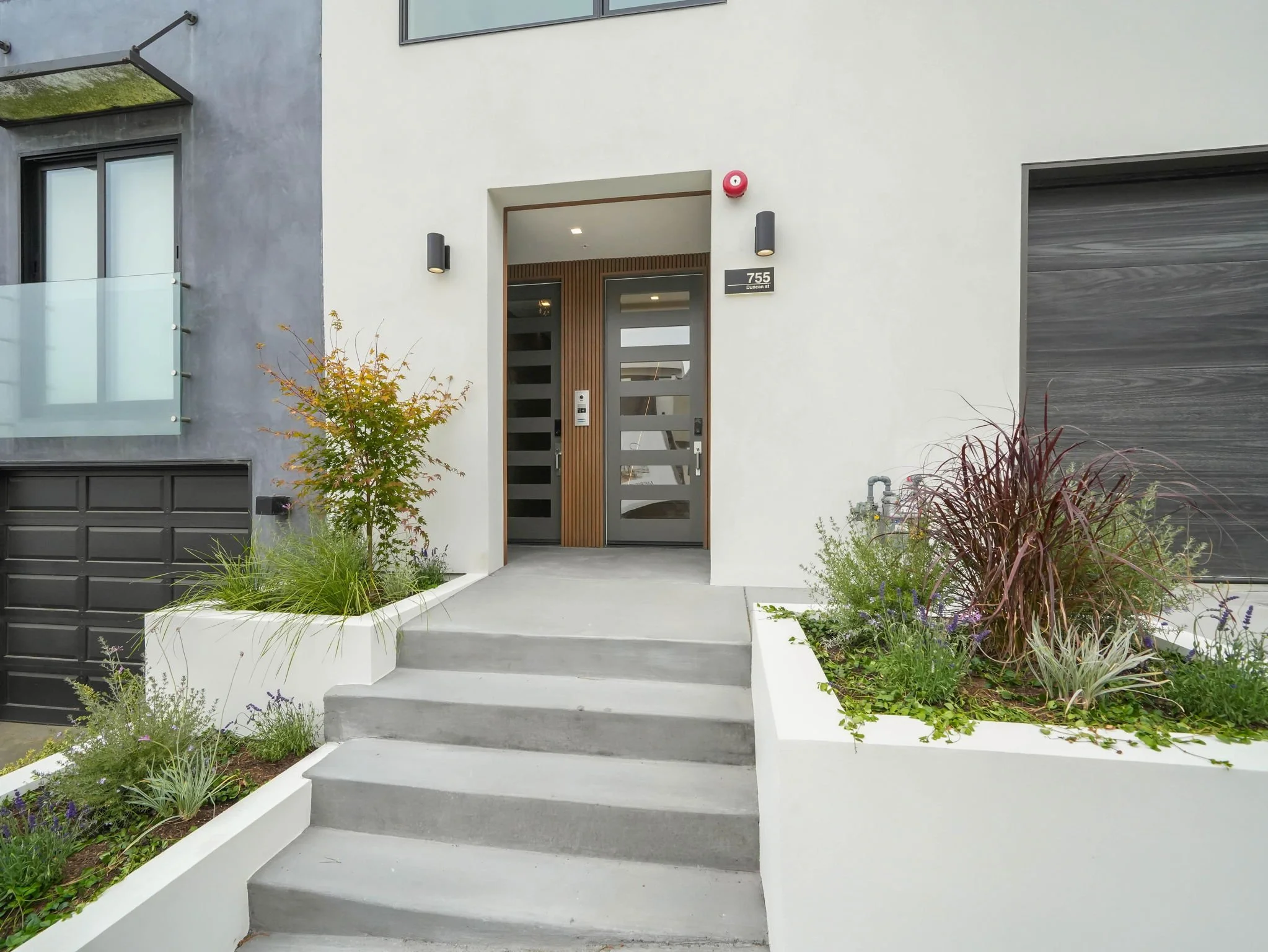 Modern residential building entrance with gray and white exterior walls, black and gray front door, and black wall sconces. Small landscaped garden with various plants and shrubs, including a small tree, in white planters, along with steps leading to