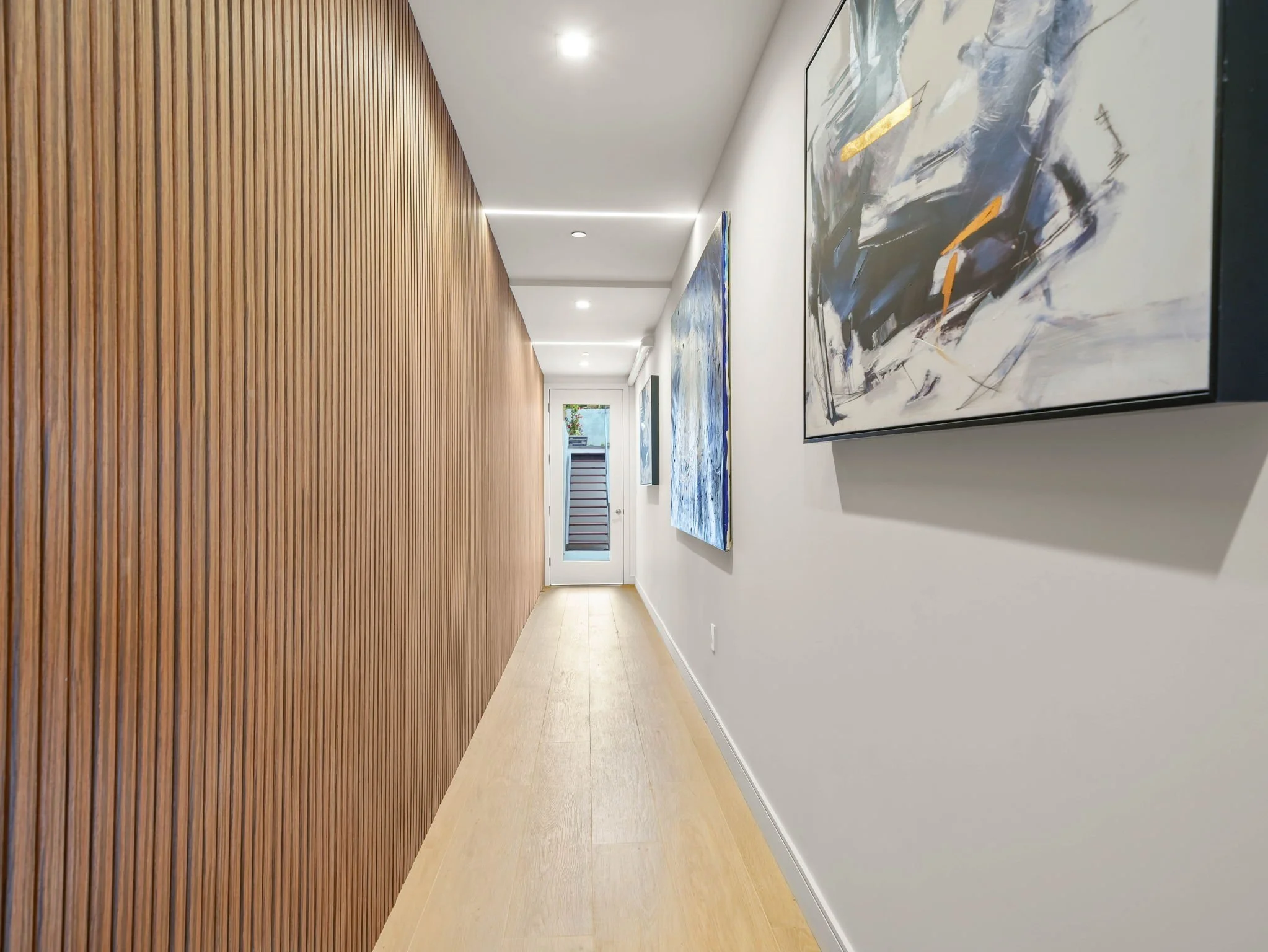 Long hallway with wooden slat wall on the left and white wall with abstract paintings on the right. Recessed ceiling lights and a door at the end of the hallway.