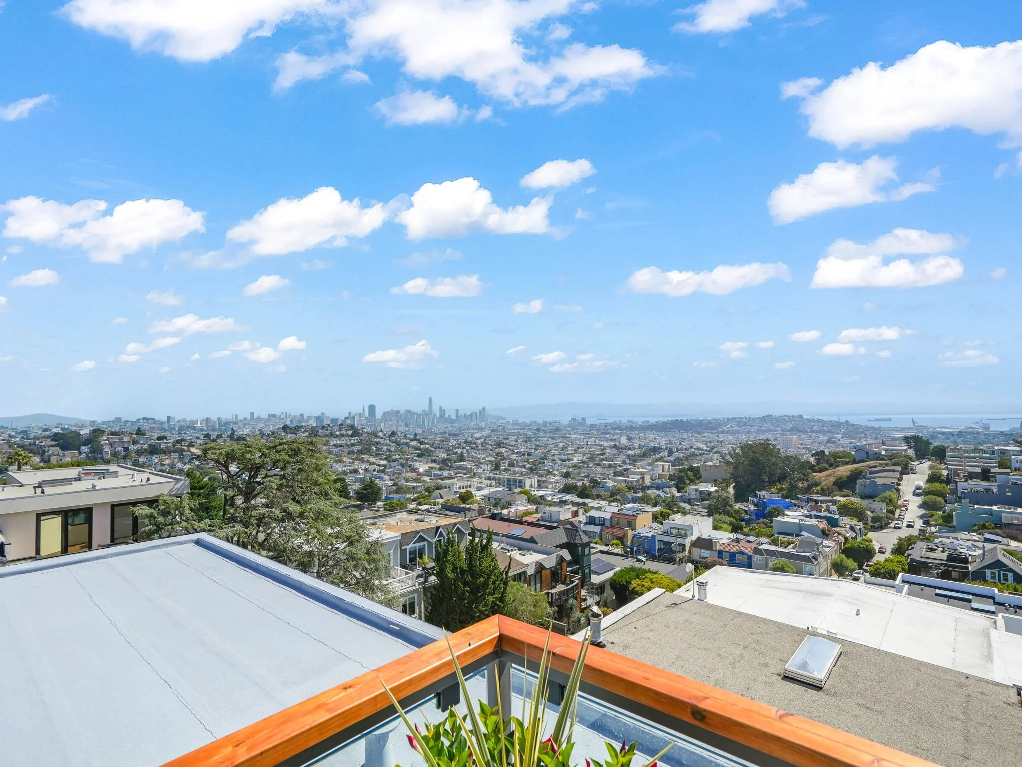 View of a city skyline from a rooftop with a modern balcony, overlooking residential neighborhoods, high-rise buildings, and a cloudy blue sky.