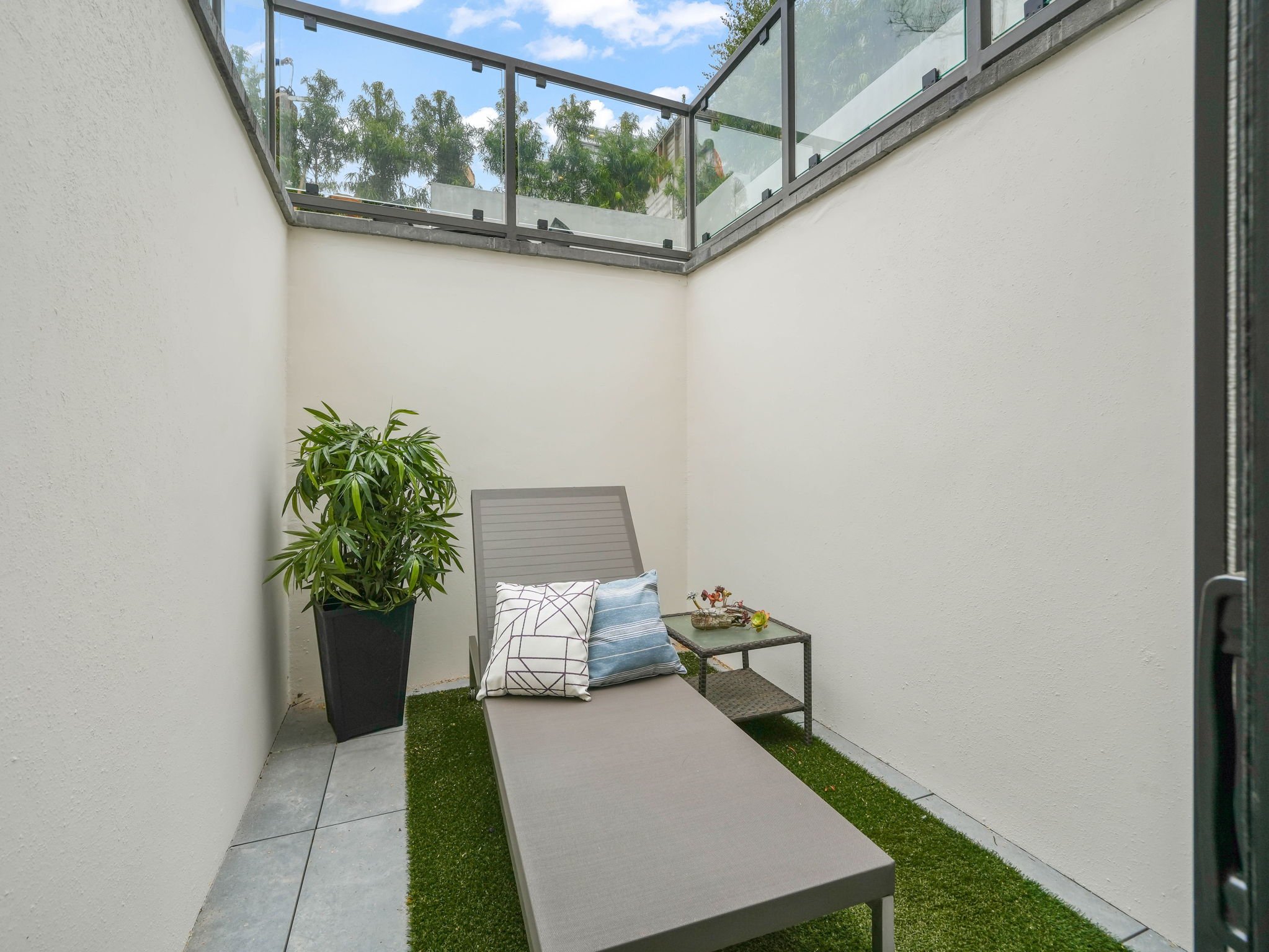 A small outdoor patio with a white wall, a large potted plant, a lounge chair with pillows, and a glass railing above. There are trees and a blue sky with clouds visible in the background.