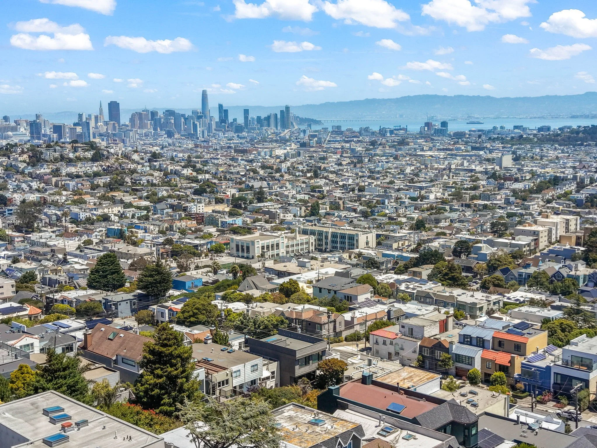 An aerial view of San Francisco cityscape, featuring numerous residential houses in the foreground and the downtown skyline with tall skyscrapers in the background, with a bridge crossing a body of water and hills in the distance under a partly cloud
