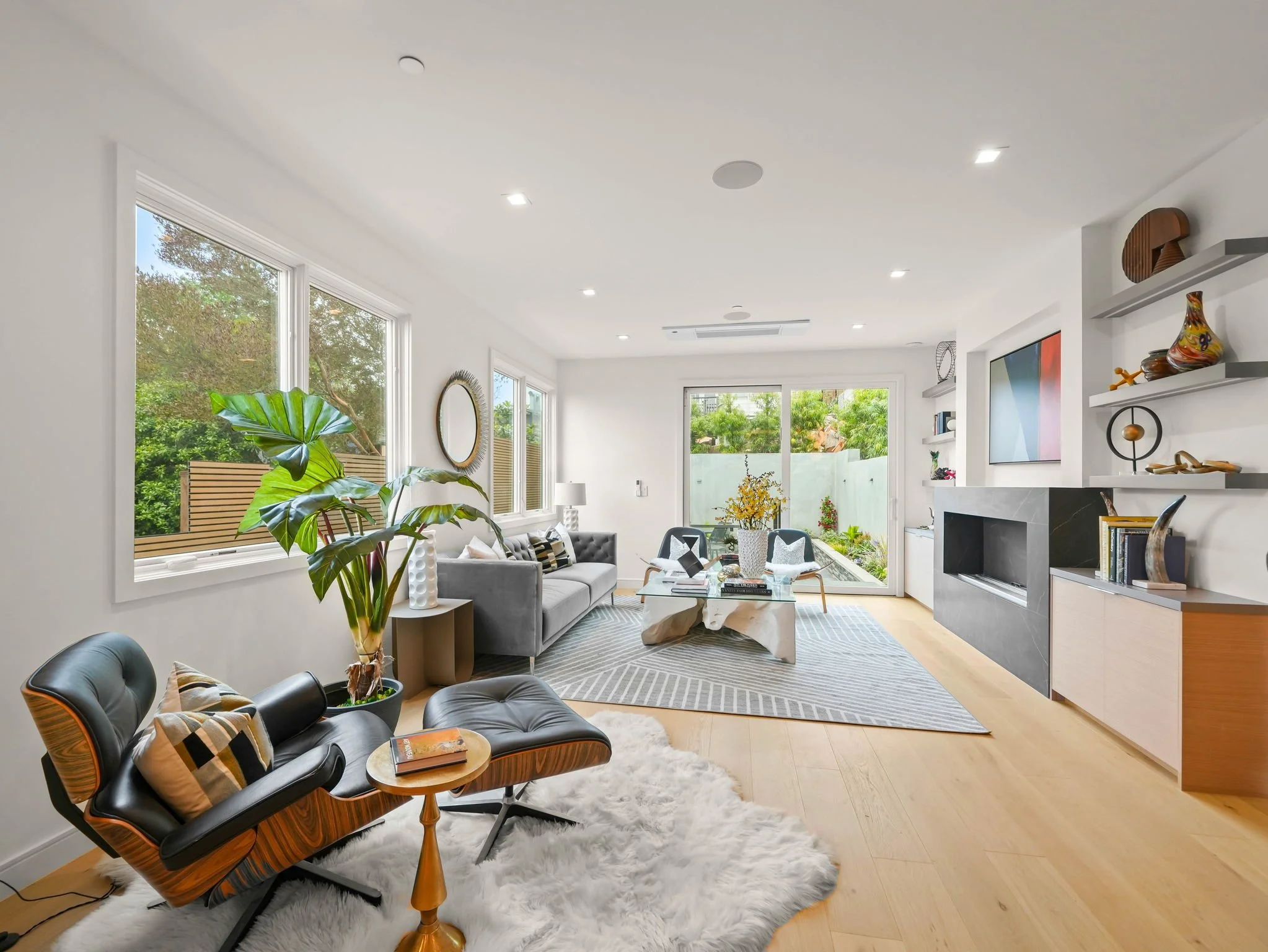 A bright, modern living room with large windows, white walls, a glass coffee table, black and wood lounge chairs with pillows, a gray sofa, a large flat-screen TV, built-in shelves with decorative objects, and indoor plants.