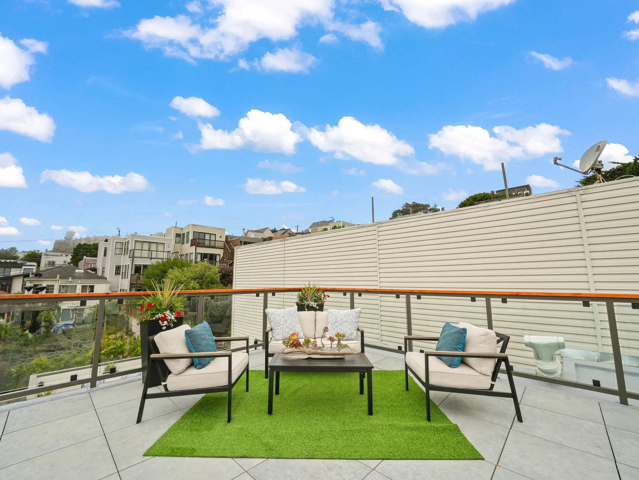 Outdoor rooftop patio with seating area, including white cushioned chairs, a black coffee table, and potted plants, enclosed by a white privacy fence under a partly cloudy sky. Apartments and houses are visible in the background.