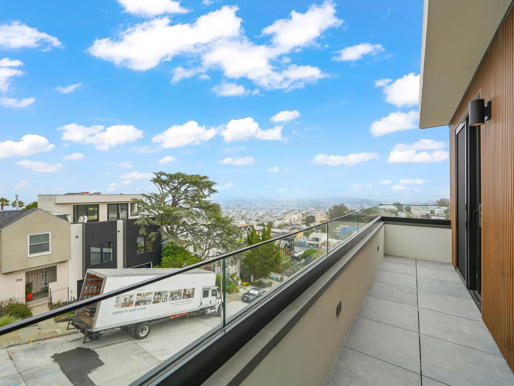 View from a balcony showing a neighborhood with modern houses, a tree, and a delivery truck parked on the street, under a bright blue sky with scattered clouds.