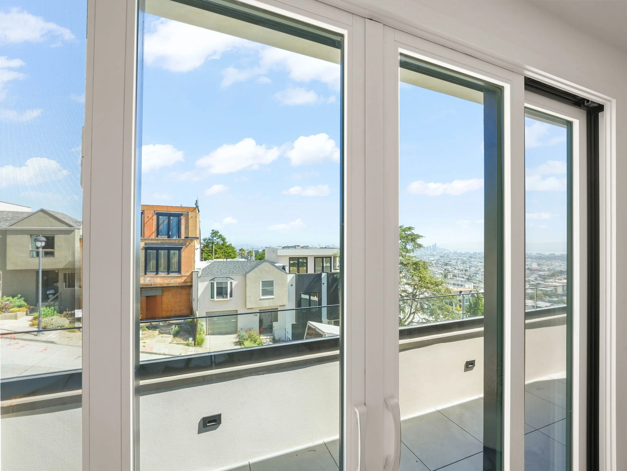 A view of a residential neighborhood and city skyline through large glass windows and a sliding glass door from inside a modern apartment or condo.