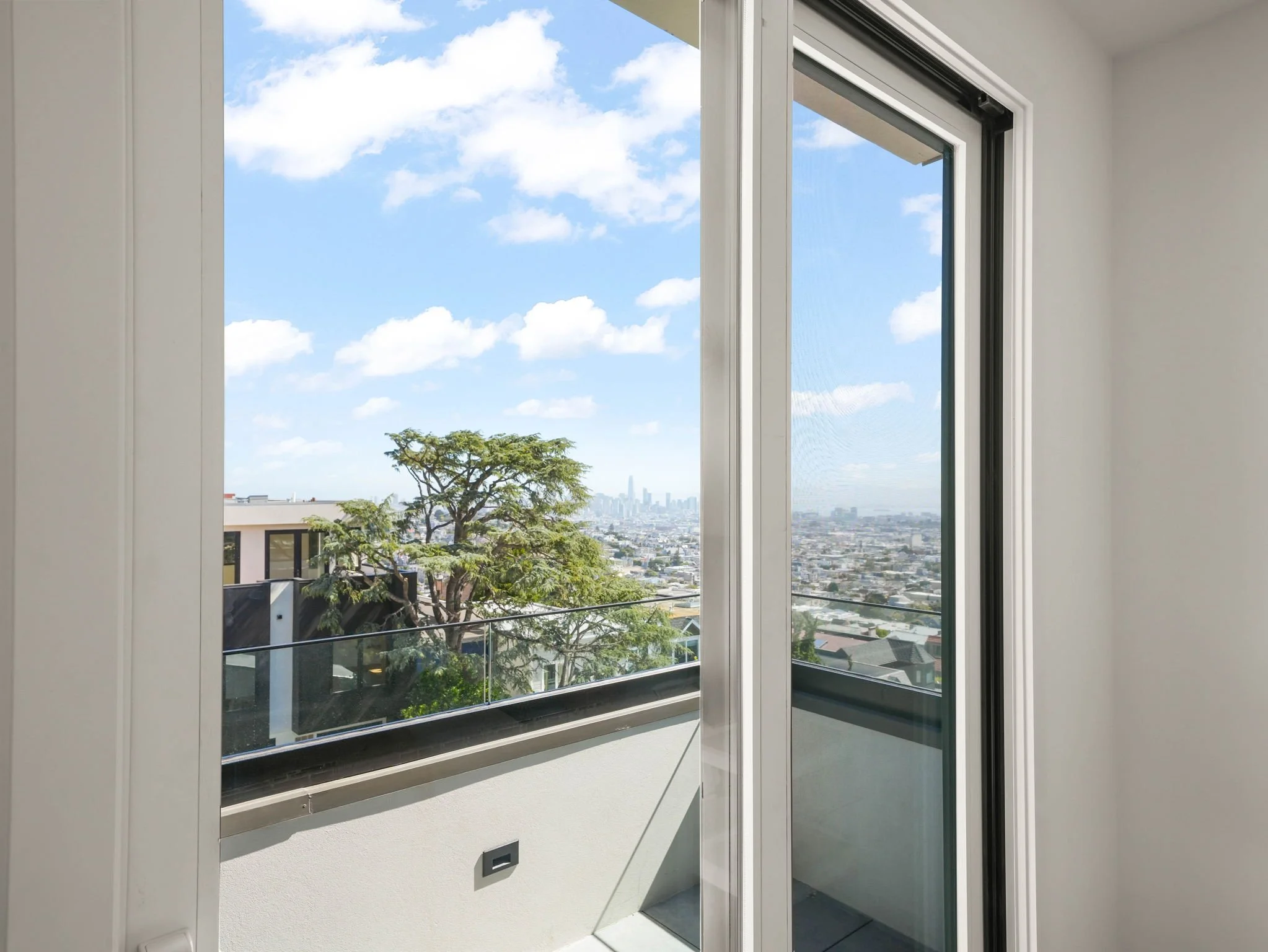 Interior view of an open sliding glass door leading to a small balcony with a cityscape and blue sky with clouds in the background.
