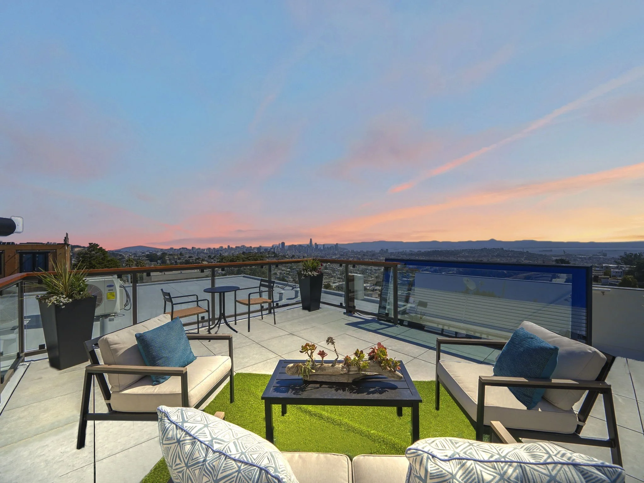 A rooftop patio with outdoor furniture, potted plants, and a view of a city skyline at sunset with pink and blue clouds.