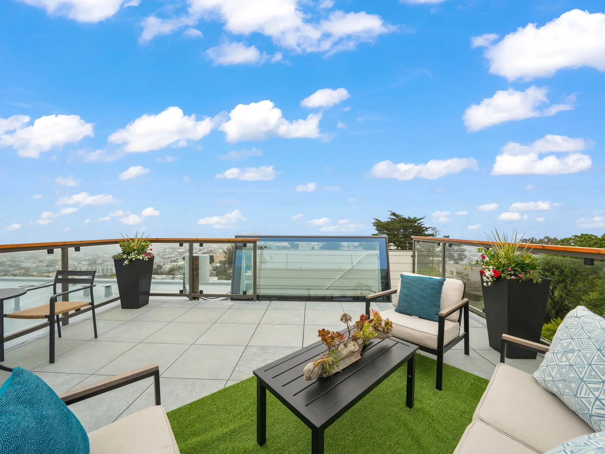 Rooftop terrace with outdoor seating, potted plants, a glass railing, and a view of the sky with scattered clouds.