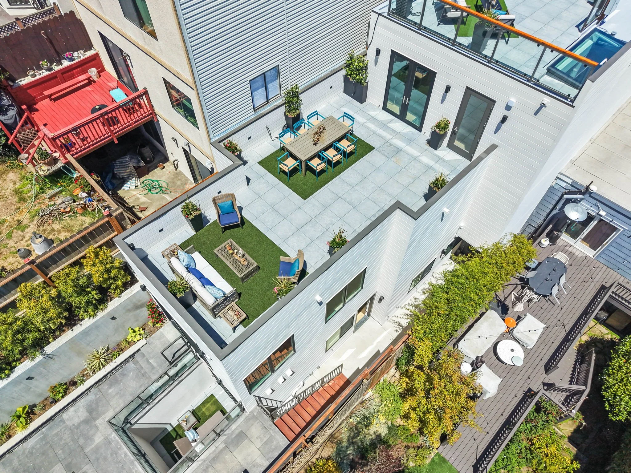 Aerial view of a modern residential building with a rooftop patio, outdoor dining table, chairs, potted plants, and surrounding greenery.