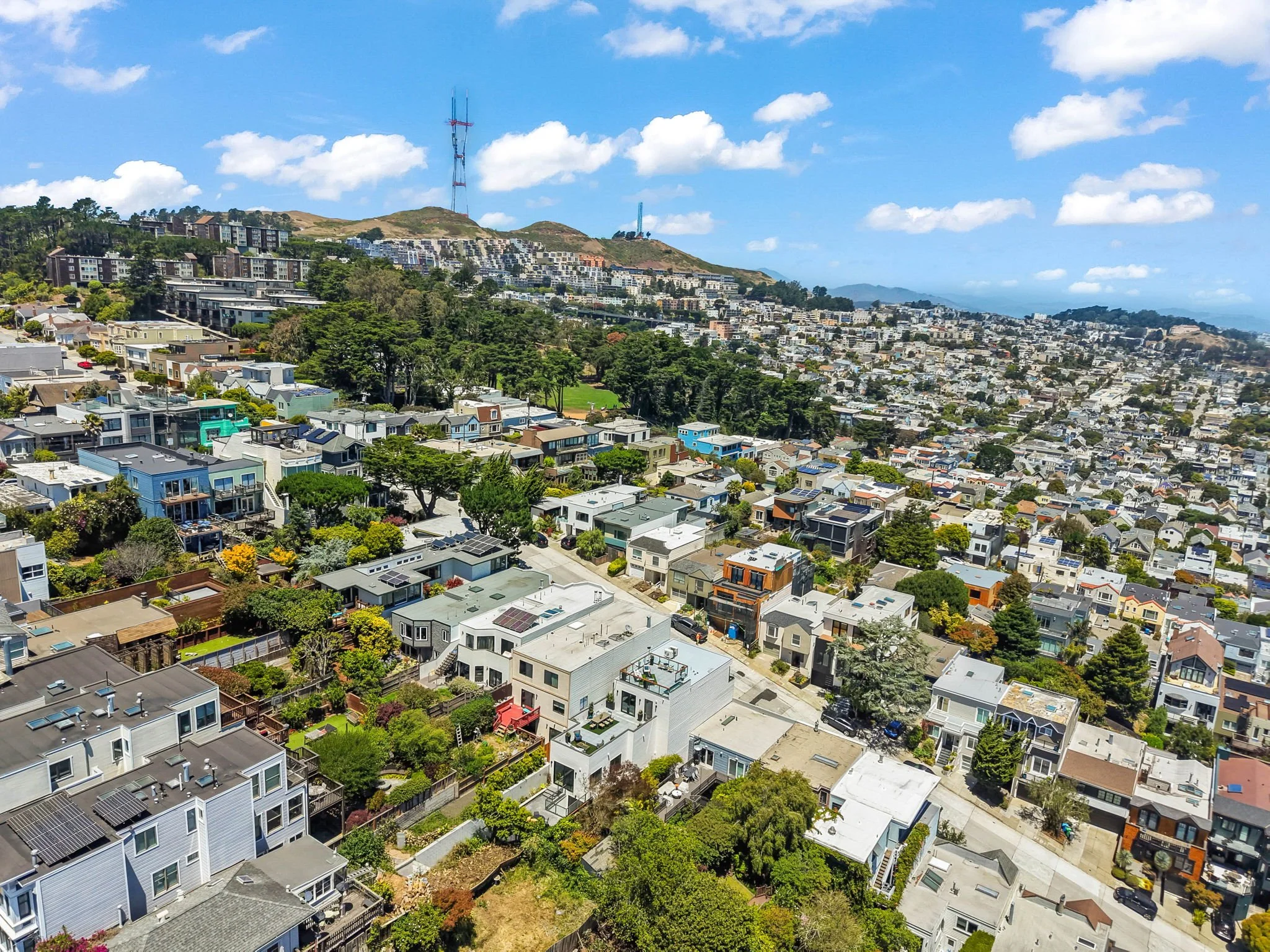 Aerial view of a hillside neighborhood with numerous houses, trees, and a large hill in the background under a partly cloudy blue sky.