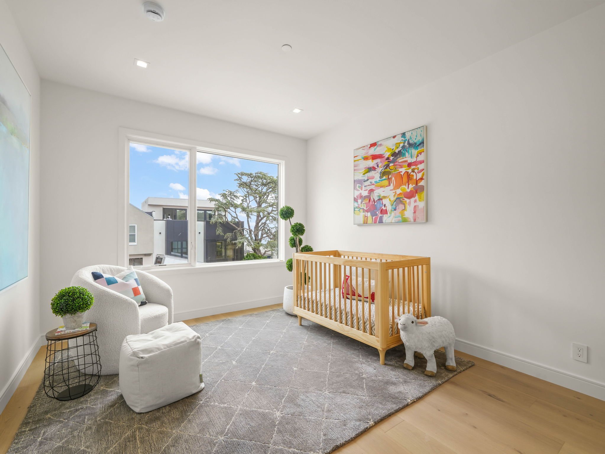 A bright nursery with white walls and a large window overlooking a tree and modern building. The room features a white armchair with colorful pillows, a wooden crib, a plush sheep toy, a small round side table with a plant, a potted topiary, a colorf