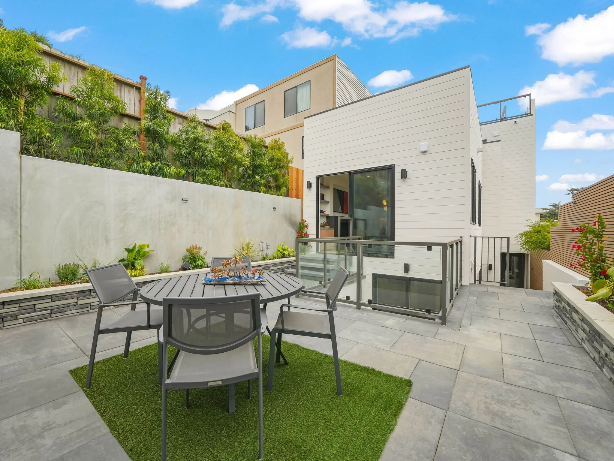 Modern white residential building with a rooftop deck, black-framed windows, and a patio with outdoor dining furniture, green plants, and a small patch of artificial grass under a bright blue sky with scattered clouds.