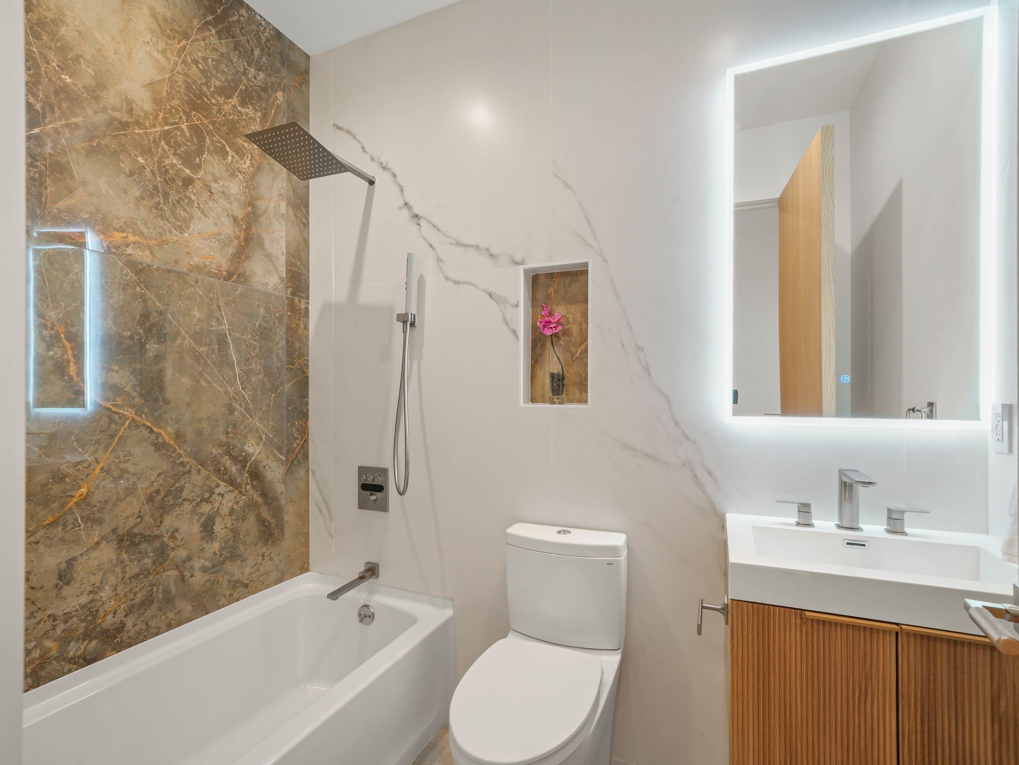 Modern bathroom with marble accent wall, walk-in shower with a rain shower head, white fixtures, large rectangular mirror with backlit LED lighting, and a wooden vanity cabinet.