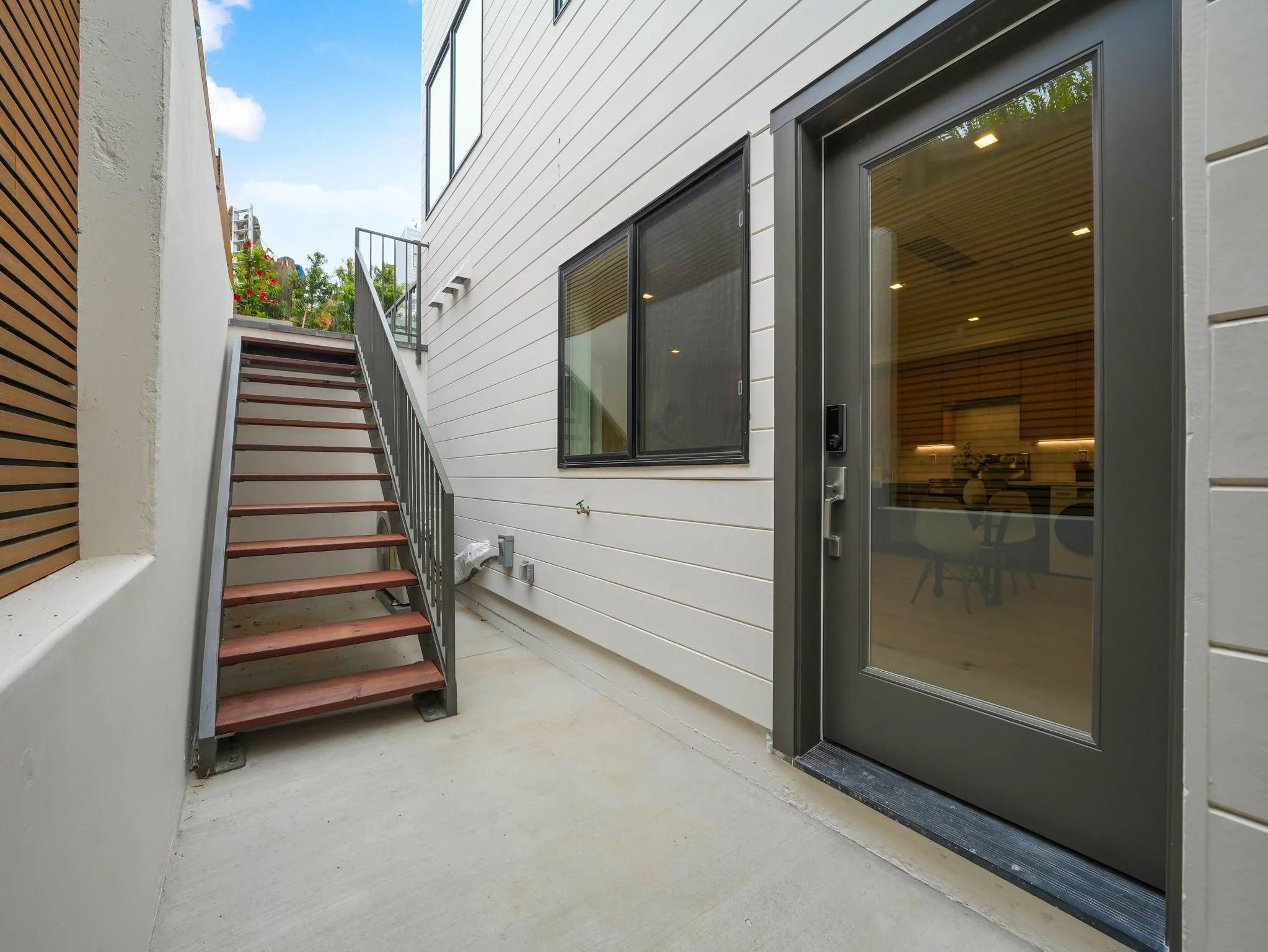 A modern outdoor patio area with concrete flooring, a staircase, and a large glass door. The building has white siding and large windows, with a wooden fence and some greenery visible in the background. The sky is blue with a few clouds.