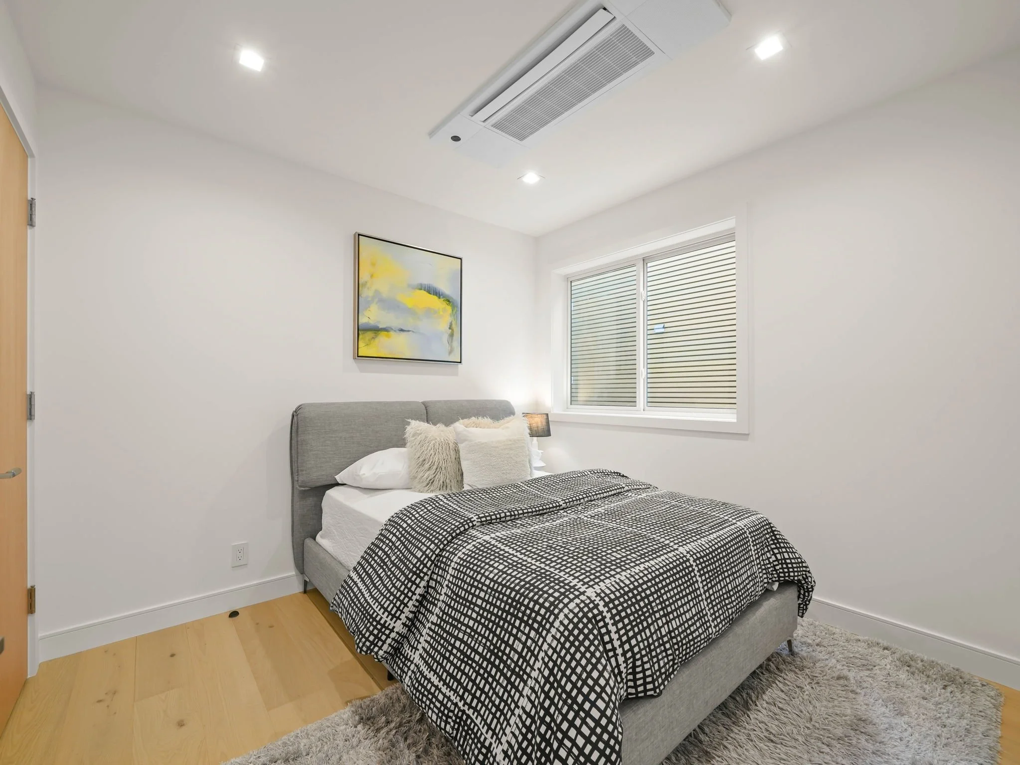 Modern bedroom with light gray upholstered bed, black and white checkered bedding, a fluffy pillows, a small lamp on bedside table, a colorful abstract painting on white wall, large window with horizontal blinds, hardwood floor, areas rug.