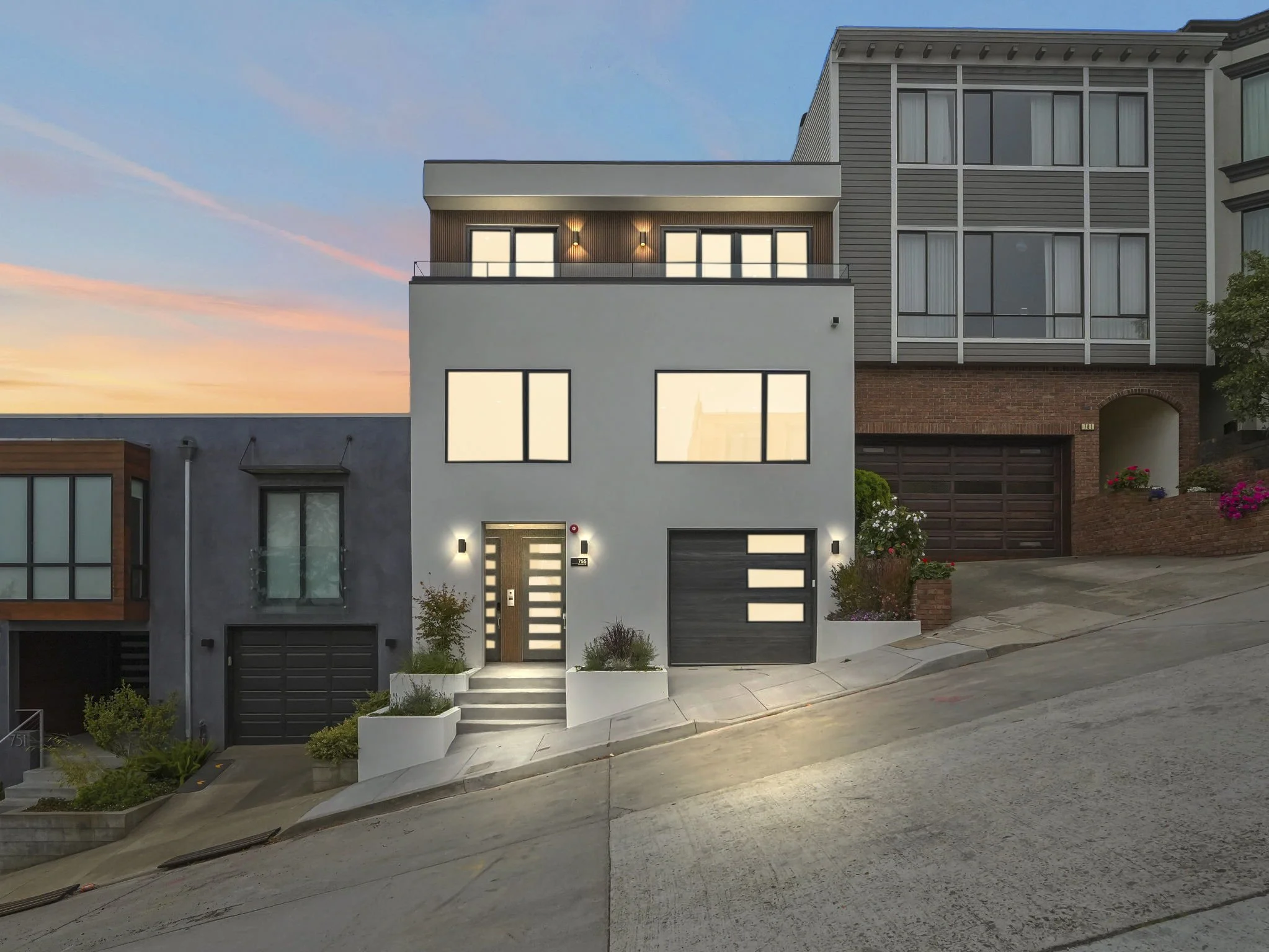 Modern three-story house with large windows, a garage, front steps, and small garden plants in a suburban neighborhood during sunset.