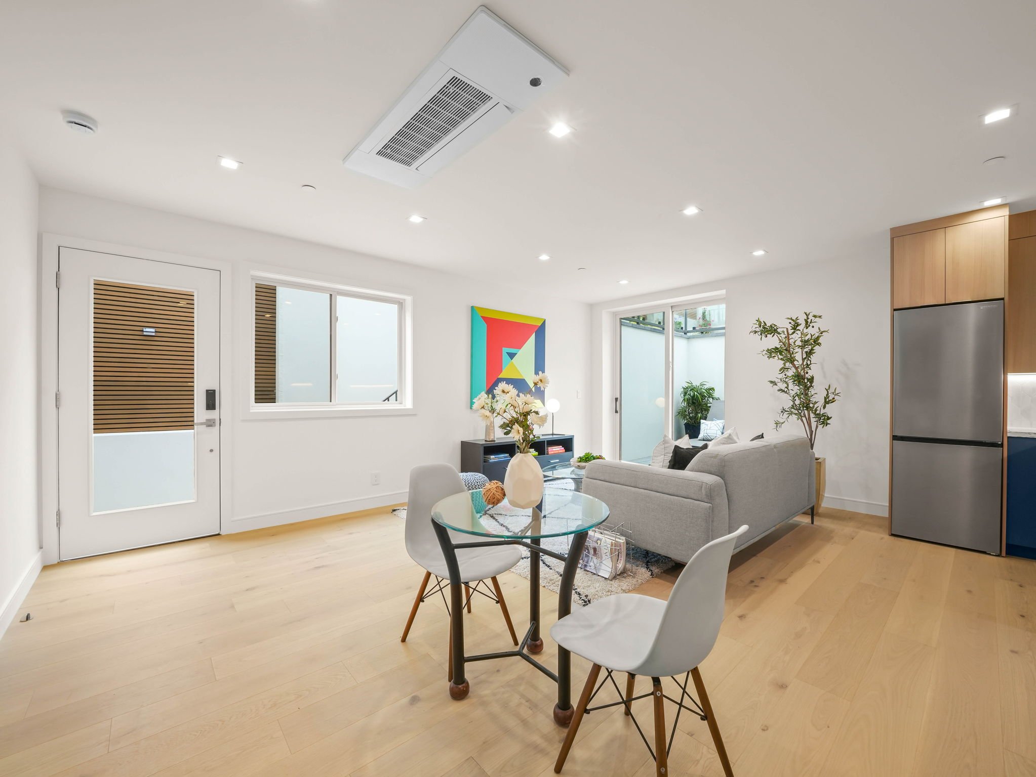 The interior of a modern living room with natural light, light wood flooring, white walls, a gray sofa, a round glass dining table with white chairs, indoor plants, artwork on the wall, and large windows.