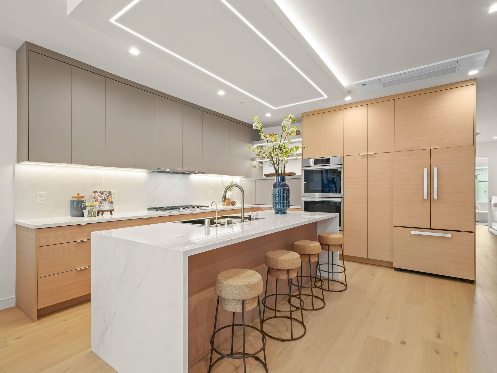 Modern kitchen with a white marble island, wooden cabinets, gray overhead cabinets, and a built-in oven. A blue vase with white flowers decorates the island. Bar stools with round cushions are lined up at the island. Recessed lighting and ceiling lig