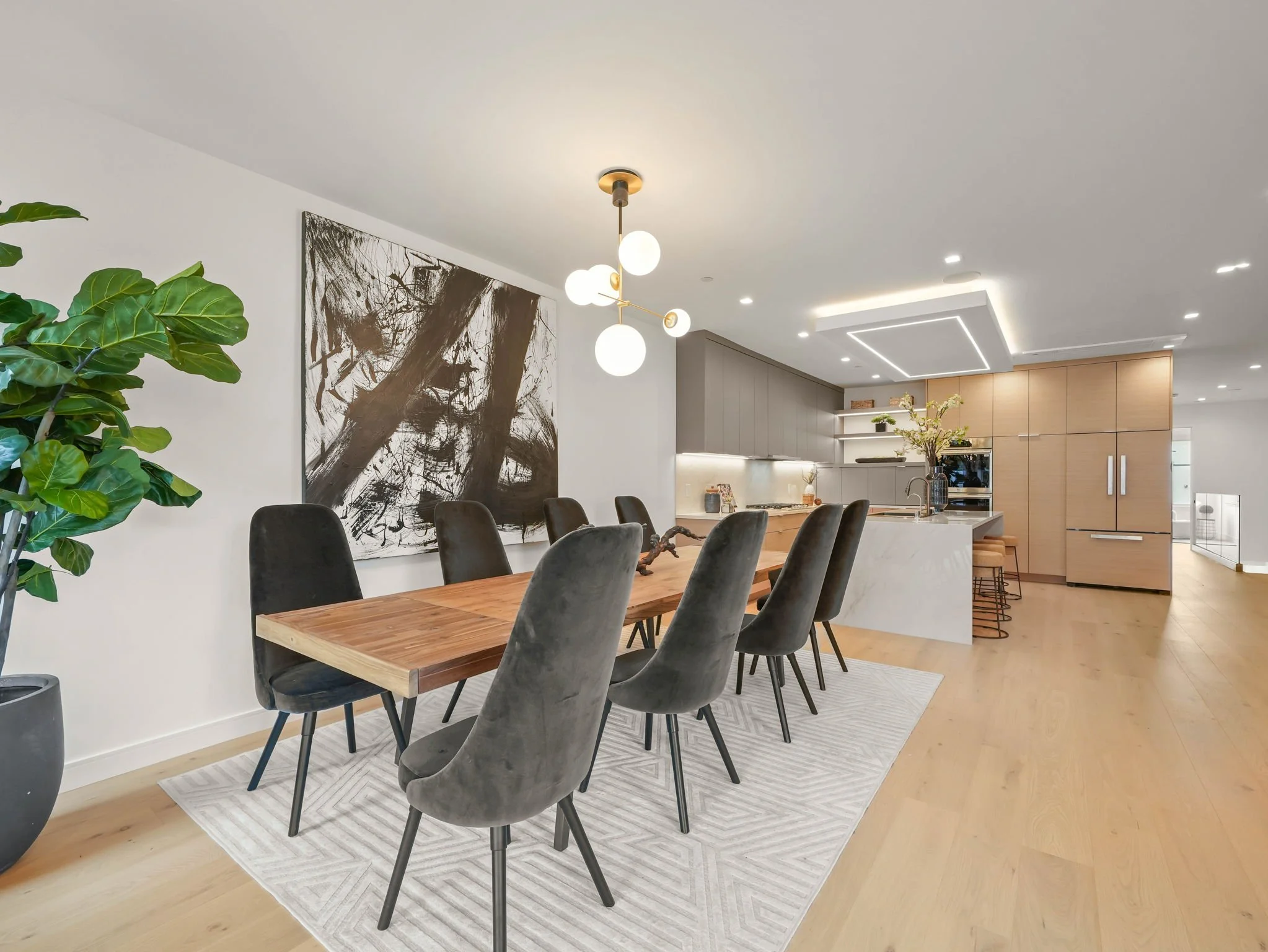 Modern dining area with a wooden table, black chairs, a large abstract black-and-white artwork, a leafy green plant, and a contemporary kitchen in the background with beige cabinetry and a white countertop.