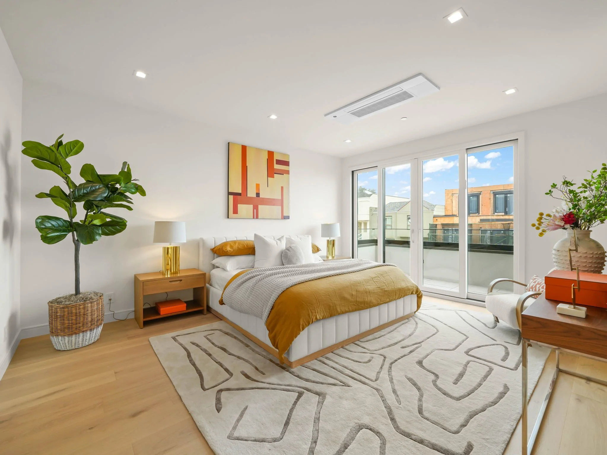 Bright living room with a white sofa, mustard cushions, a wooden side table with gold lamps, a large potted plant, colorful abstract art on the wall, and wide glass doors leading to a balcony with city buildings outside.