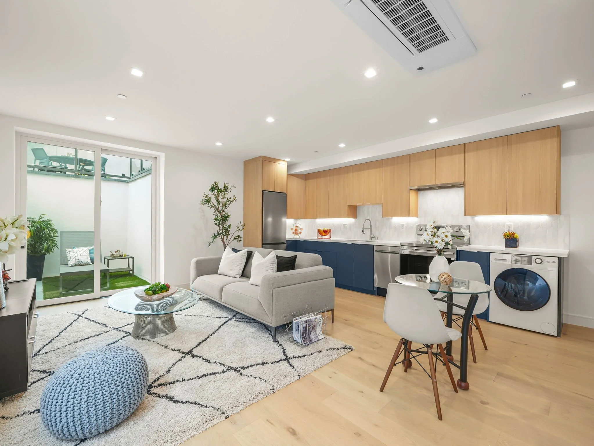 Modern open-plan living area and kitchen with white walls, wooden cabinets, and large sliding glass door opening to a small balcony or patio.