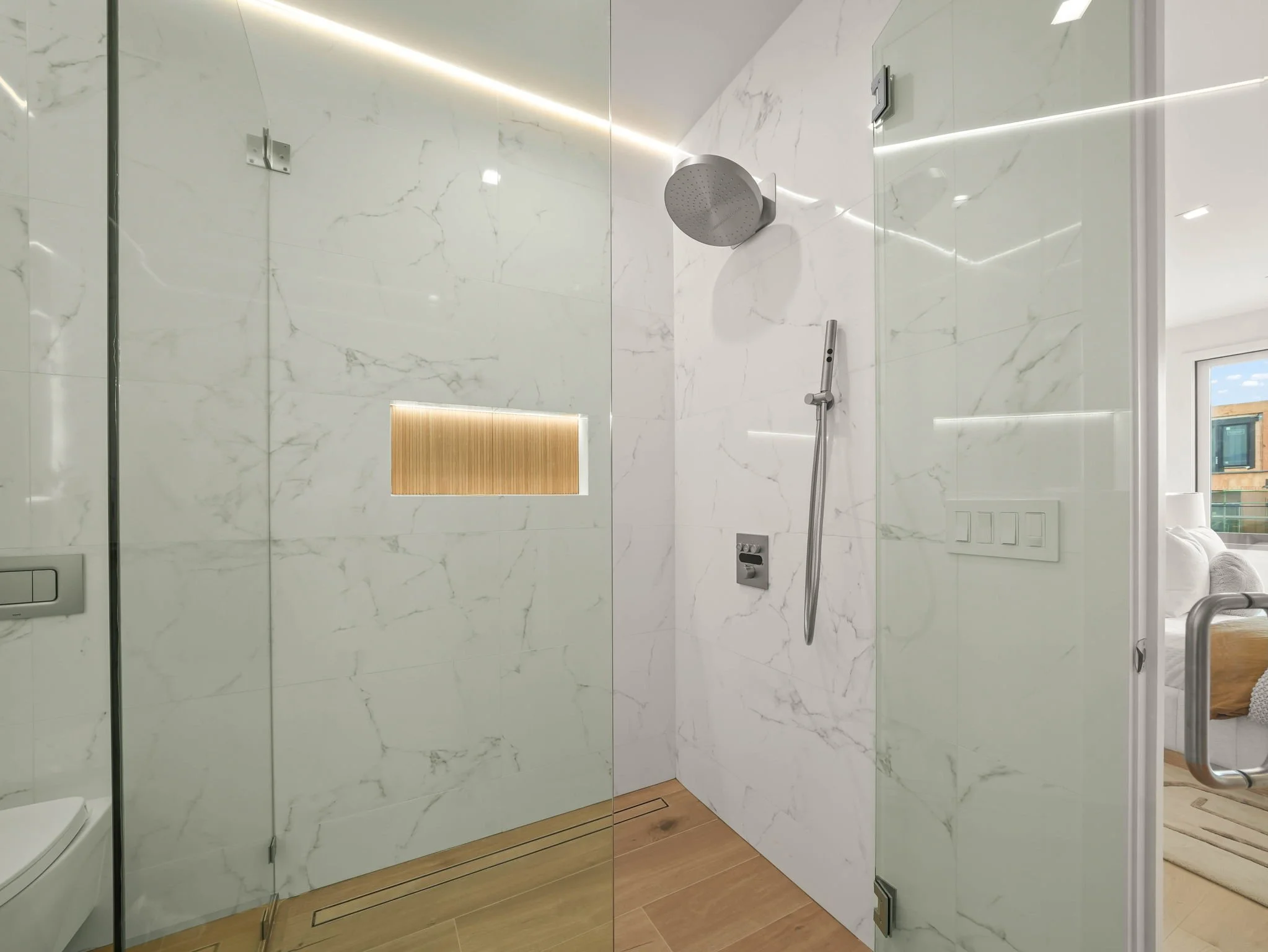 Modern shower area with white marble walls, a ceiling-mounted showerhead, a handheld shower, and wooden flooring. Reflections on a glass shower door show part of a room with a window and a glimpse of a sofa.