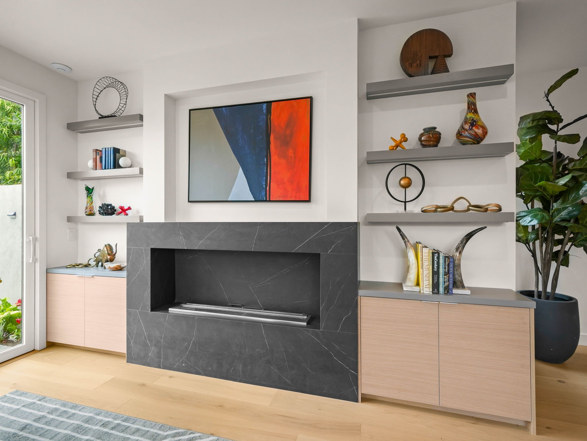 Modern living room with a gray marble fireplace, abstract painting, built-in shelves with decorative objects, and a large window with greenery outside.