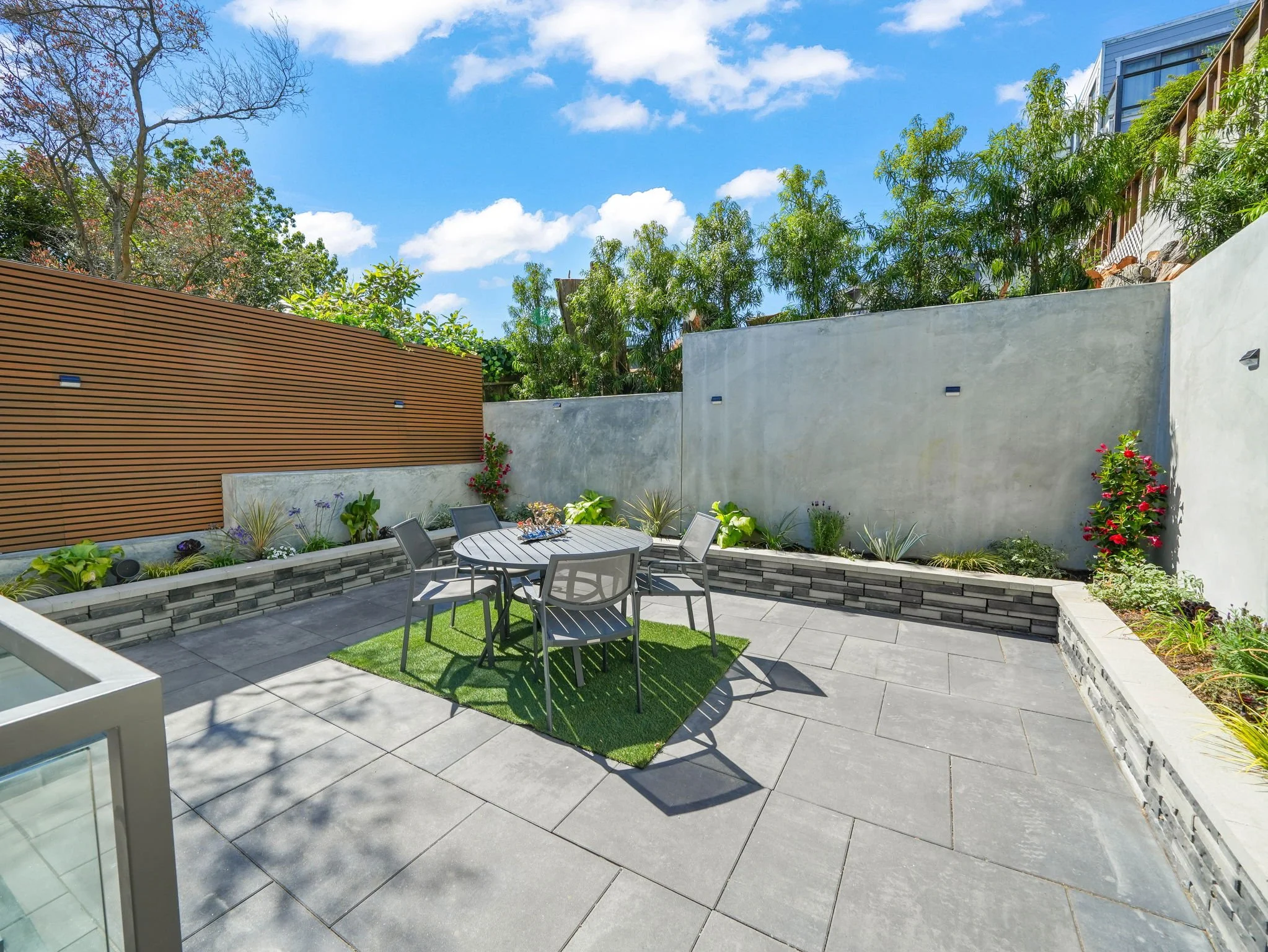 A modern backyard patio with gray tile flooring, a round dining table with chairs, a small artificial grass patch, and lush green plants along the perimeter. The sky is blue with scattered clouds, and a tall tree with bare branches is visible in the 