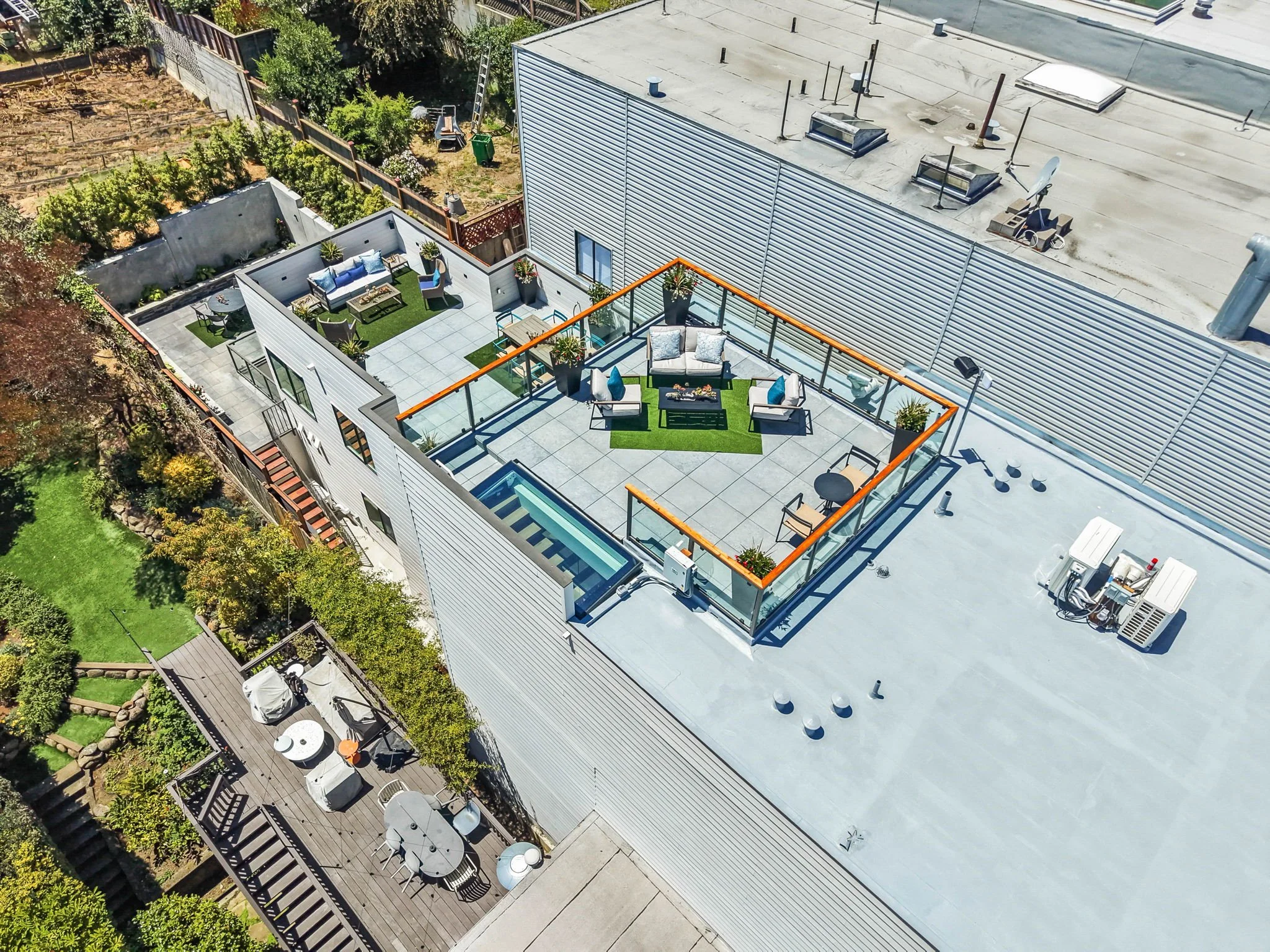 Aerial view of a rooftop patio with outdoor seating, potted plants, and a small pool, adjacent to a commercial building with a metal roof. There is a neighboring garden and yard with trees and a wooden deck.