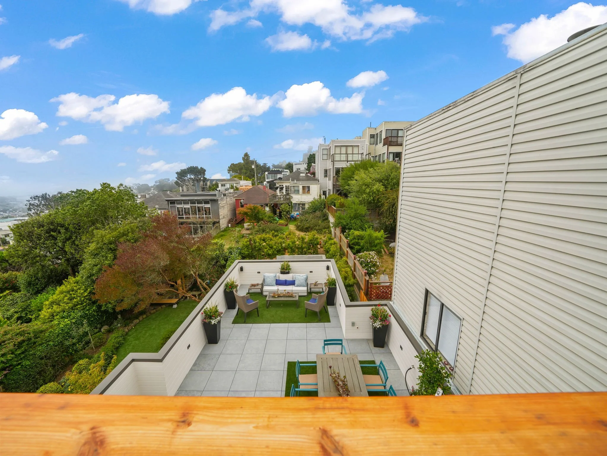 View from a balcony overlooking a backyard patio, garden, and neighboring houses under a partly cloudy sky.