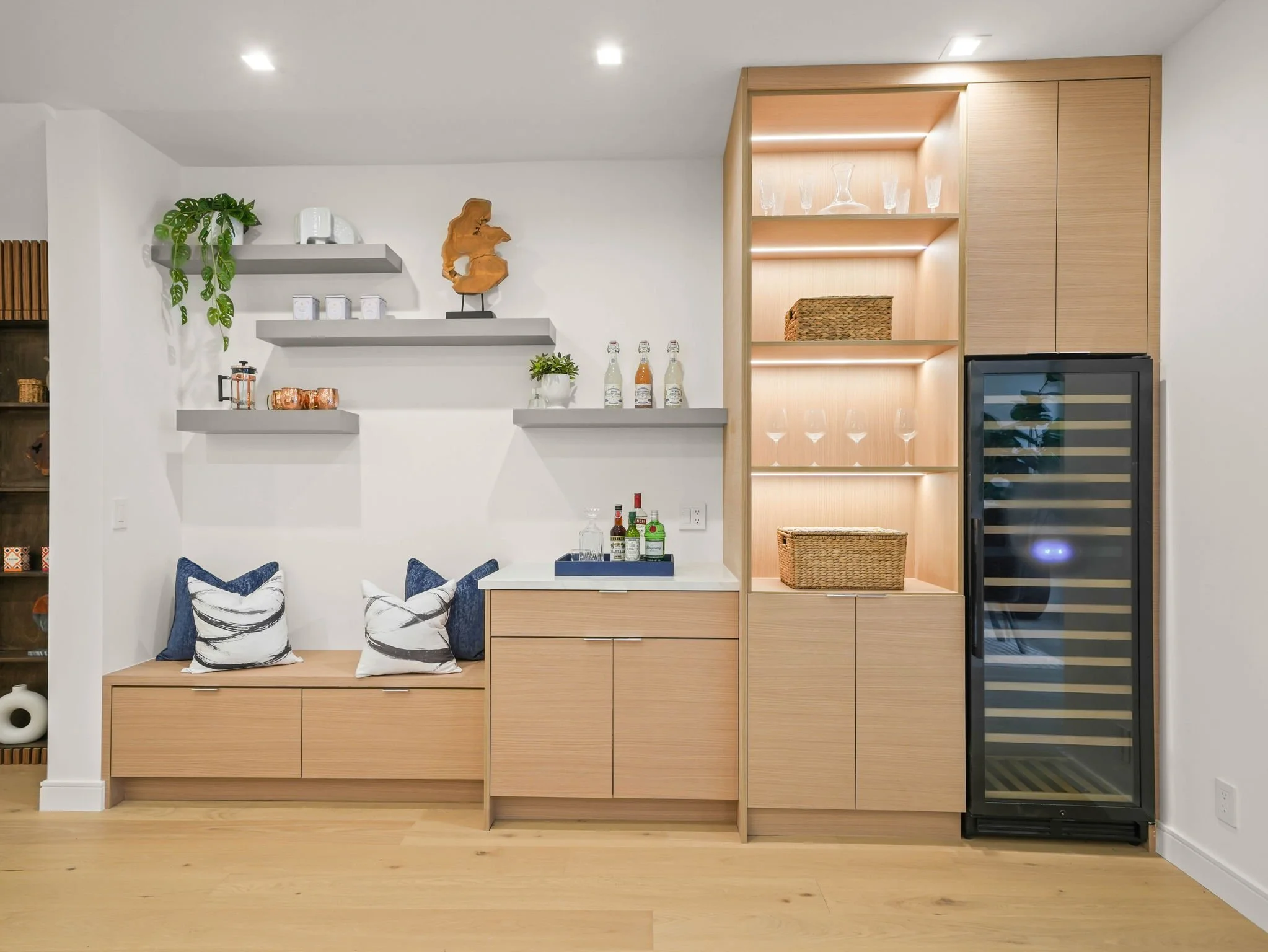 A modern kitchen with wooden cabinets, open shelving with glassware and decorative items, a built-in wine fridge, and a small seating area with pillows.