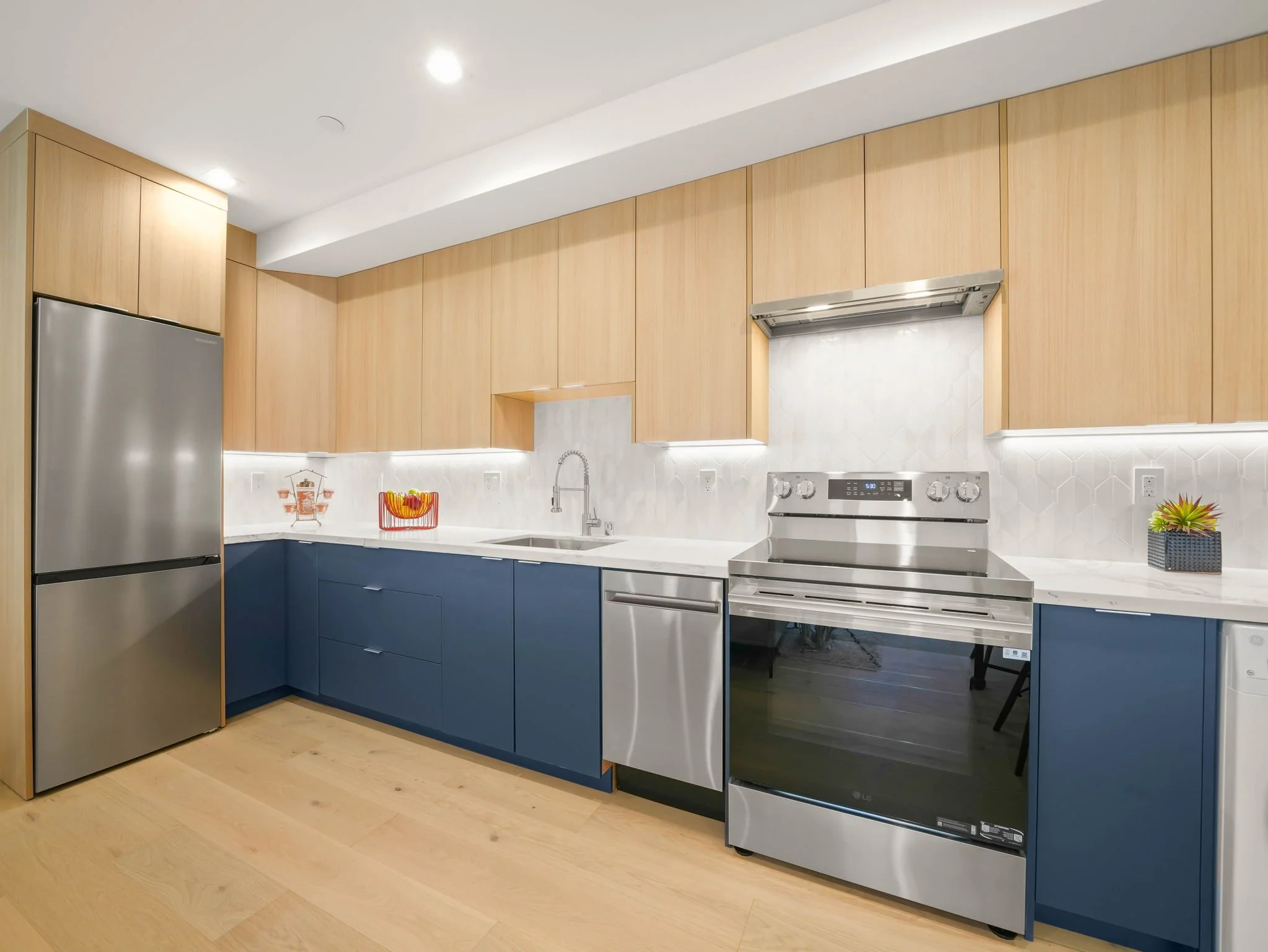 Modern kitchen with light wood cabinets, dark lower cabinets, stainless steel refrigerator, oven, dishwasher, and a black countertop with a sink, small decorative plants, and colorful utensils.