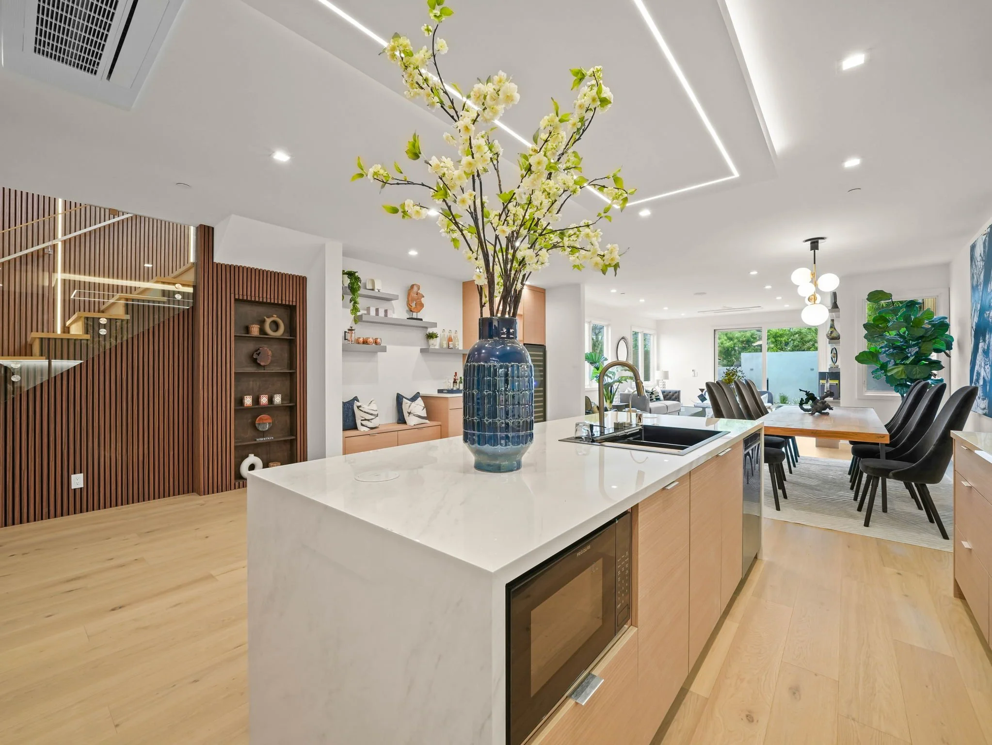 A modern open-concept kitchen and dining area with a large white island, black bar stools, a vase with white flowers on the island, built-in shelves and wood accents, and large windows in the background.