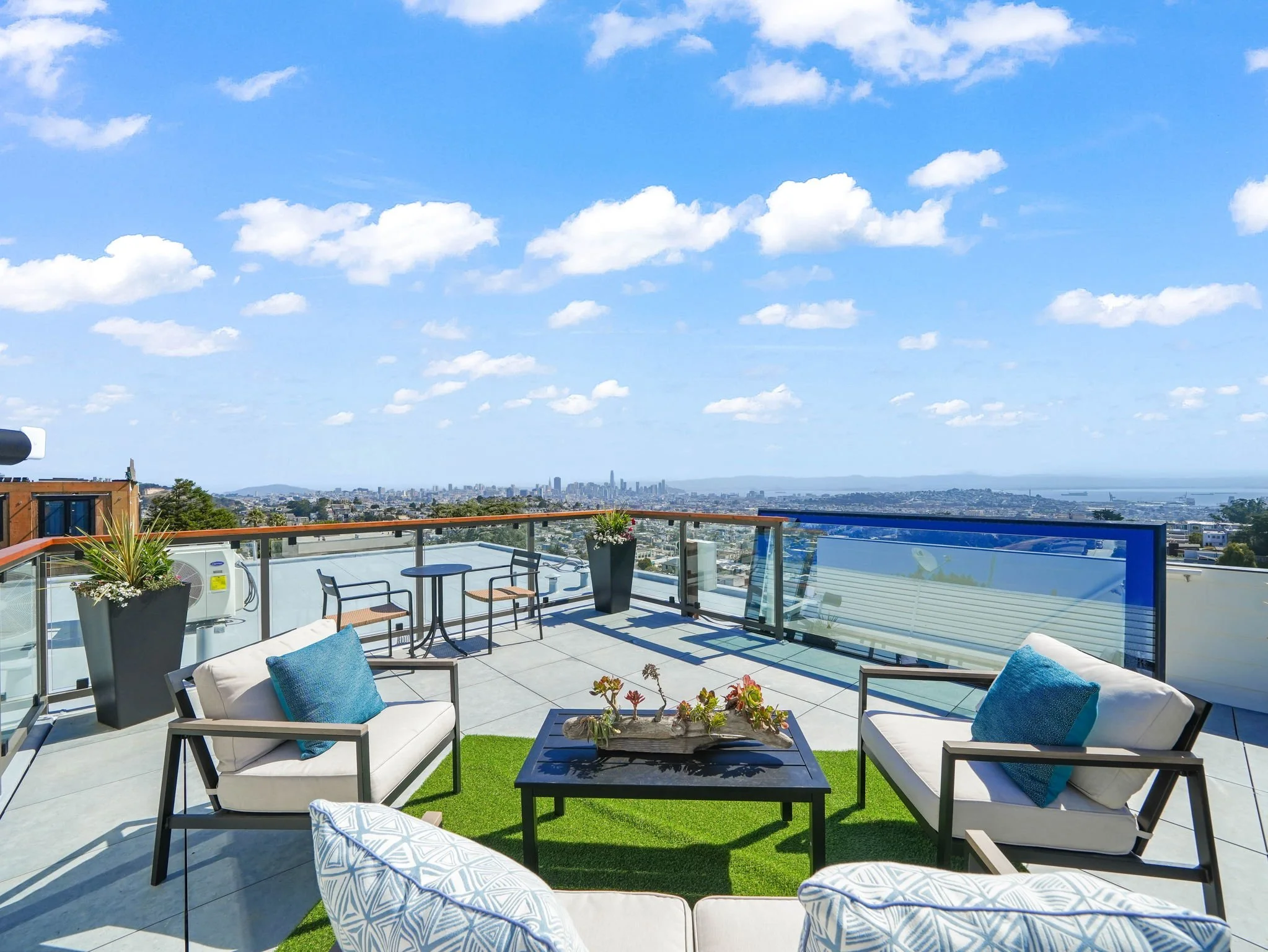View of a rooftop balcony with outdoor furniture, potted plants, and city skyline in the distance under a partly cloudy sky.