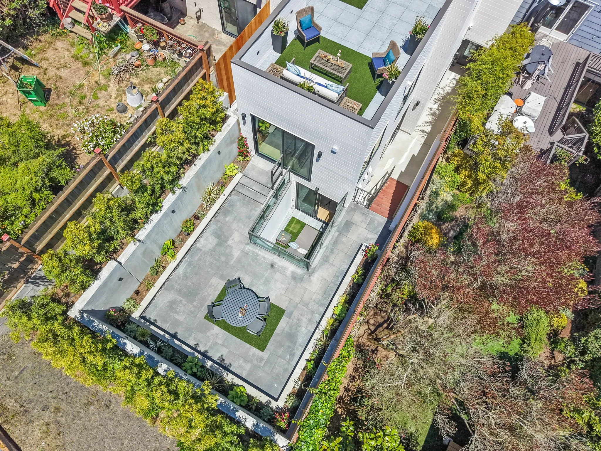 An aerial view of a modern rooftop patio with outdoor seating, a dining table with chairs, potted plants, and rooftop fencing, surrounded by trees and neighboring rooftops.