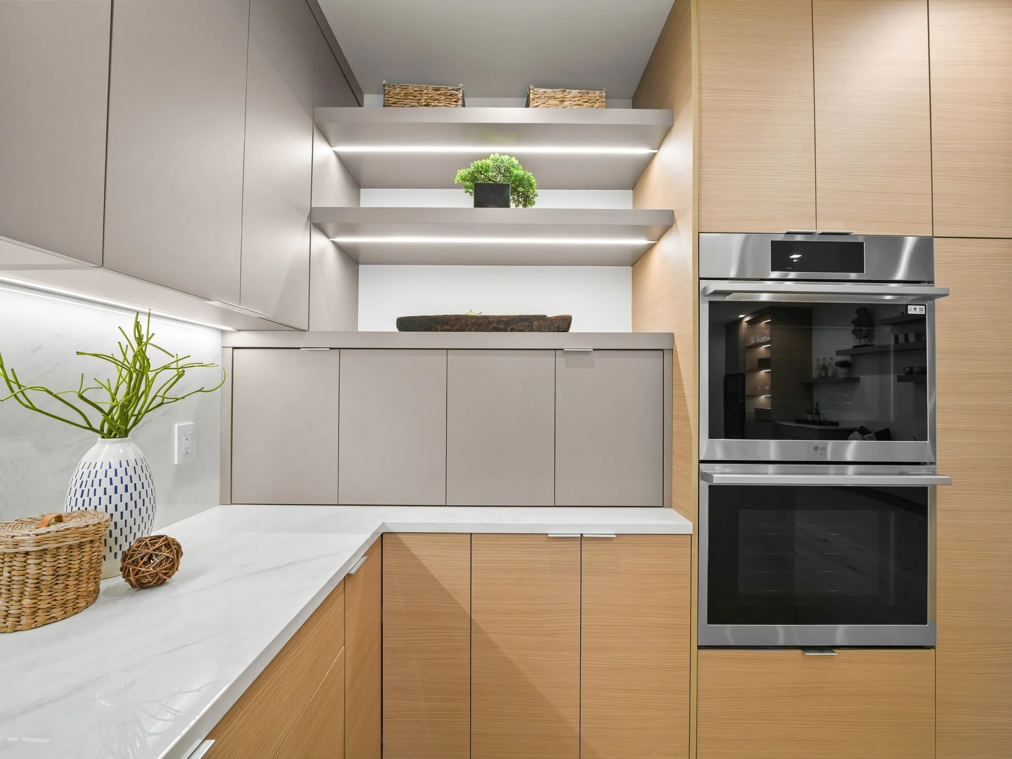Modern kitchen with wooden cabinets, stainless steel oven, white marble countertop, and decorative items including a green plant, woven baskets, and decorative balls.